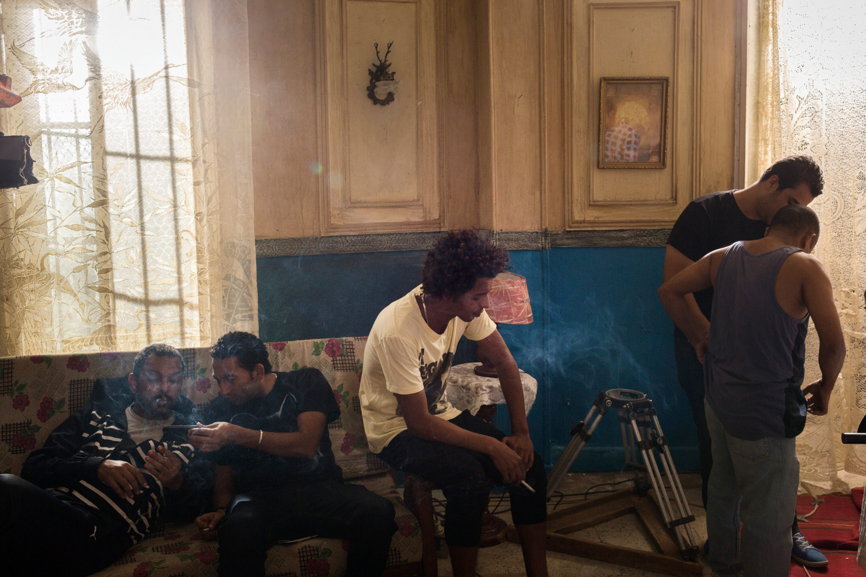 Cast and crew rest on a sofa in a sunlit room with smoke in the air and a tripod in front.