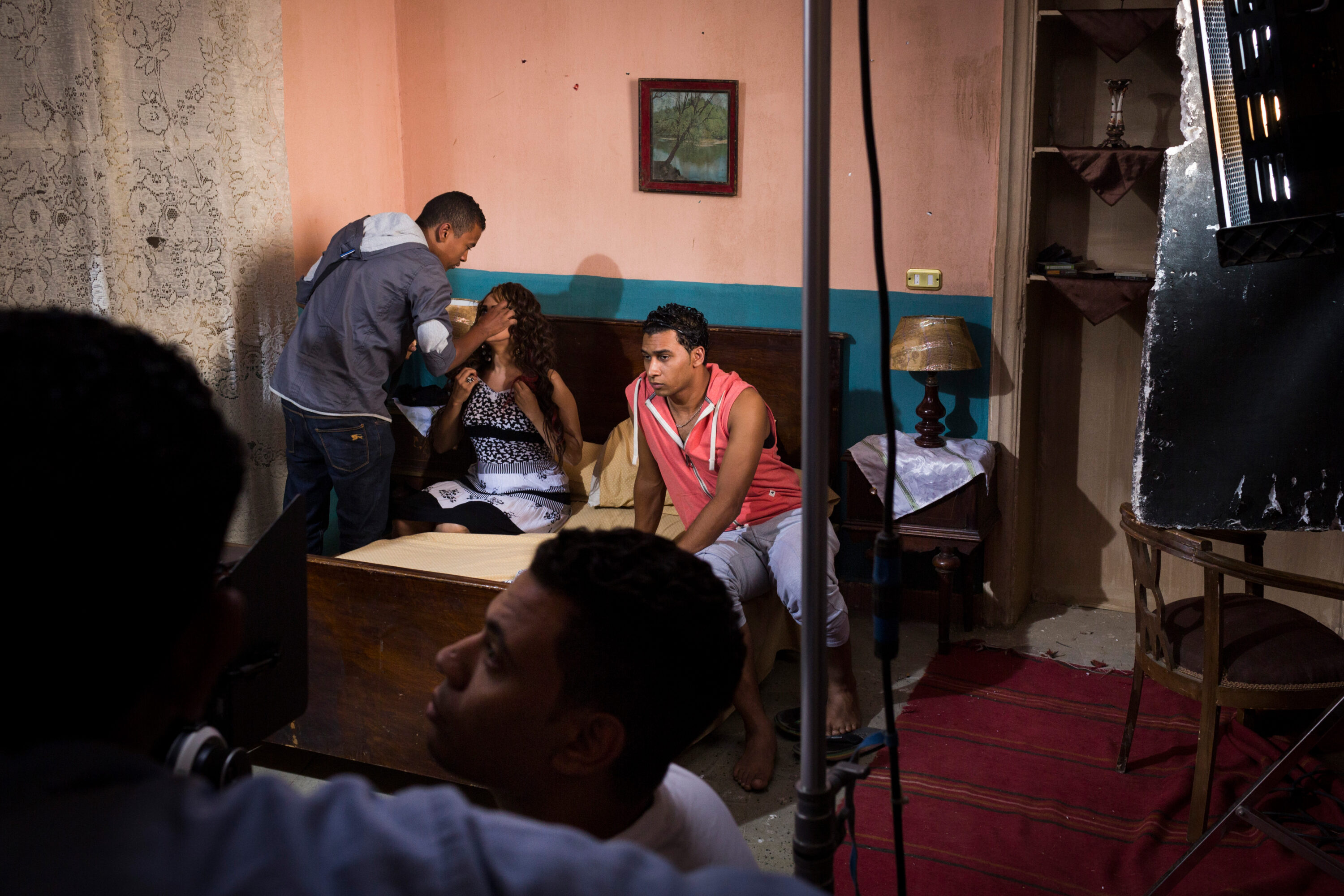 An actress has her makeup done on a bedroom set while a camera operator watches in the foreground.