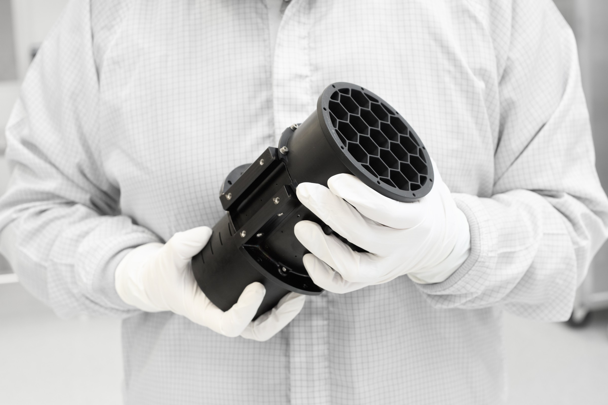 White-gloved hands holding a black cylindrical component against a white cleanroom suit at Optimax.