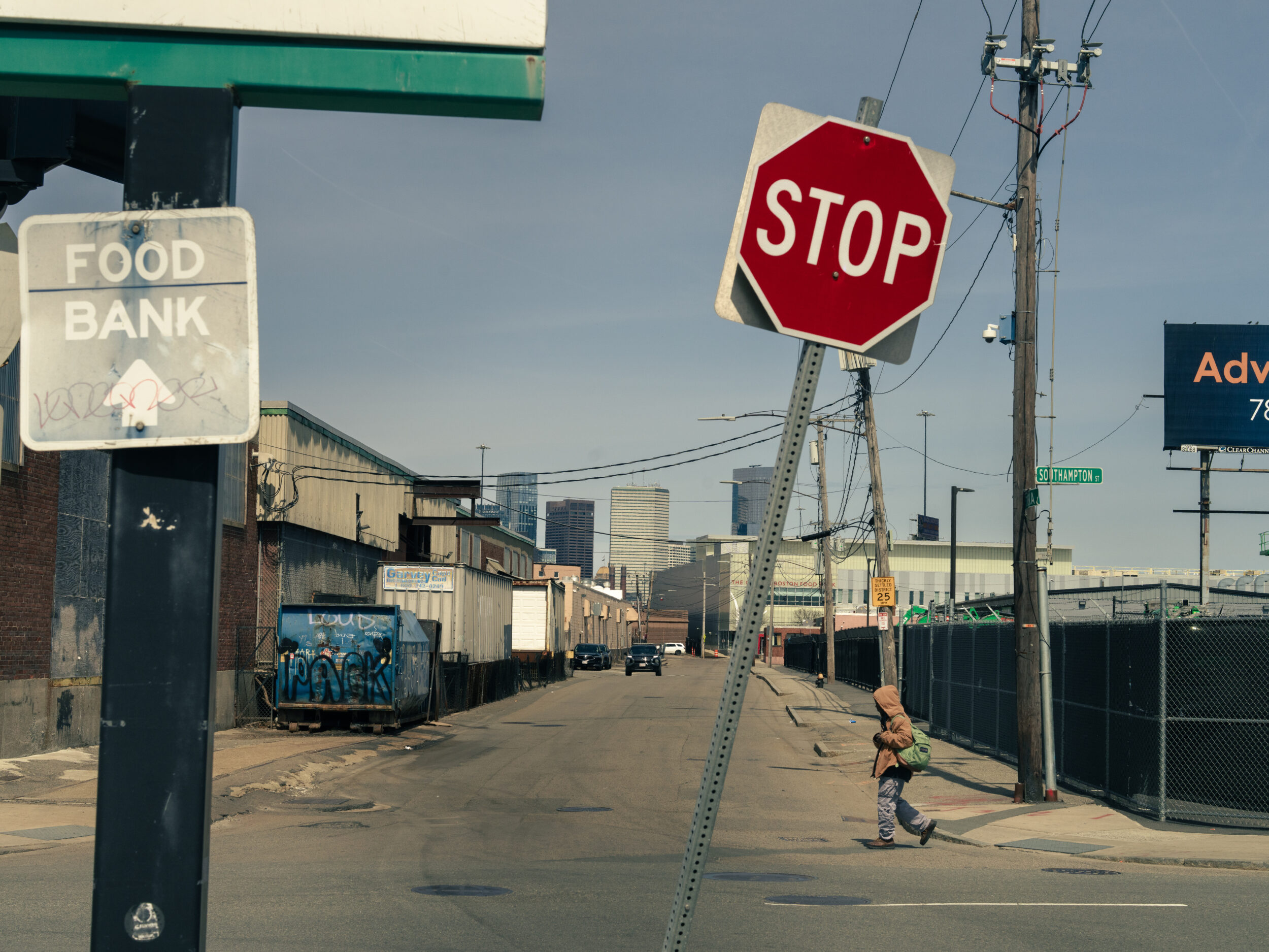 A person walks past a Food Bank sign and a leaning stop sign on Southampton Street near Mass and Cass in Boston with the city skyline in the distance A person walks past a Food Bank sign and a leaning stop sign on Southampton Street near Mass and Cass in Boston with the city skyline in the distance
