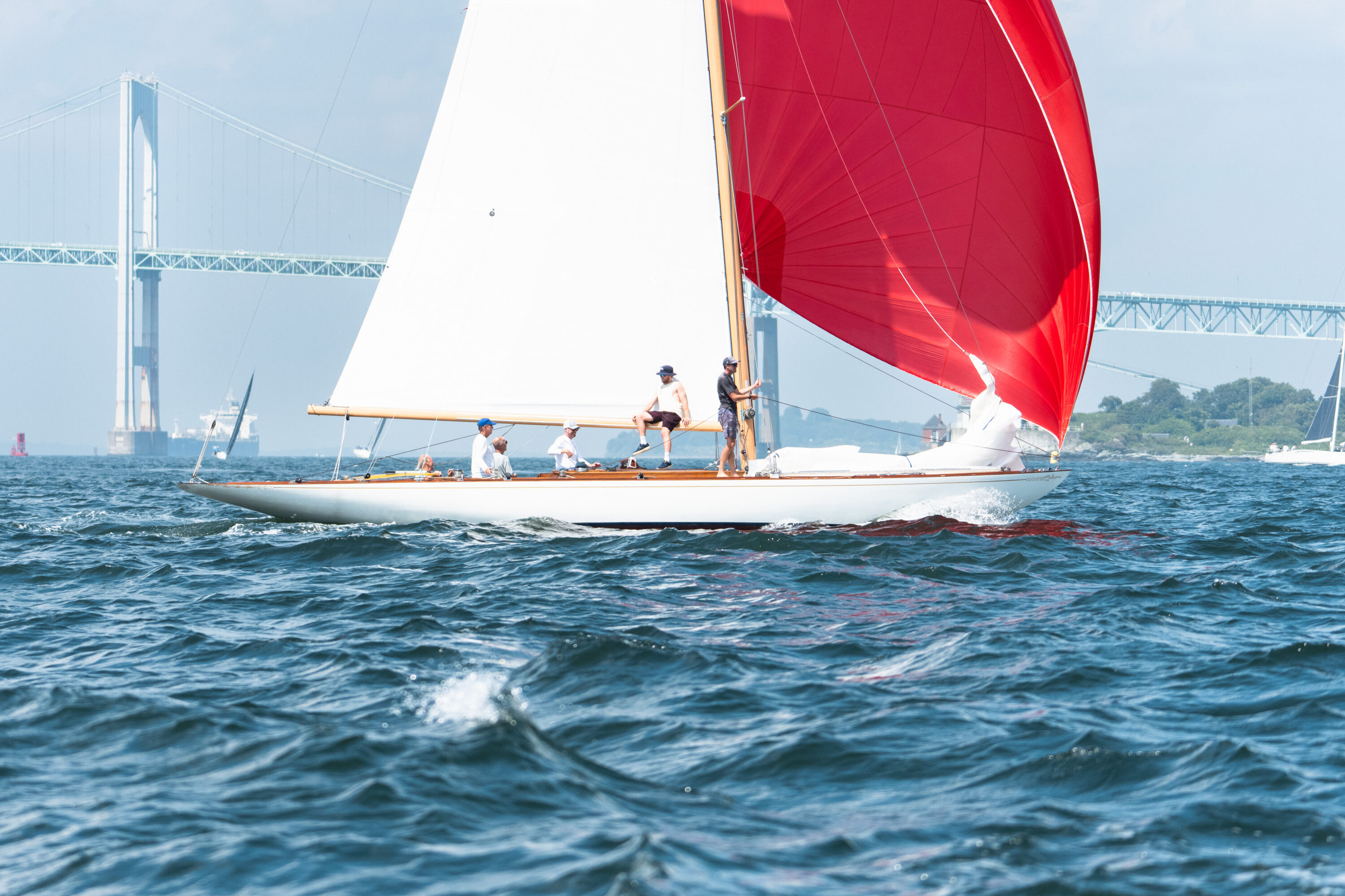 A sailboat with a billowing red spinnaker passes in front of the bridge over blue water.