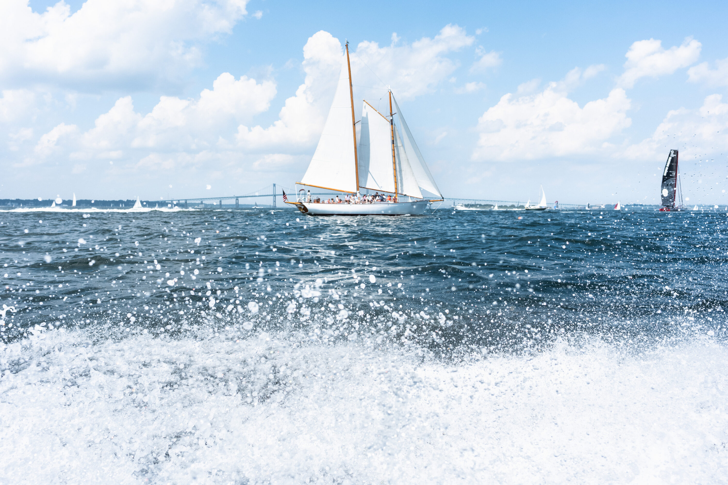 A white-sailed boat crosses the bay beyond bright spray thrown up in the foreground.