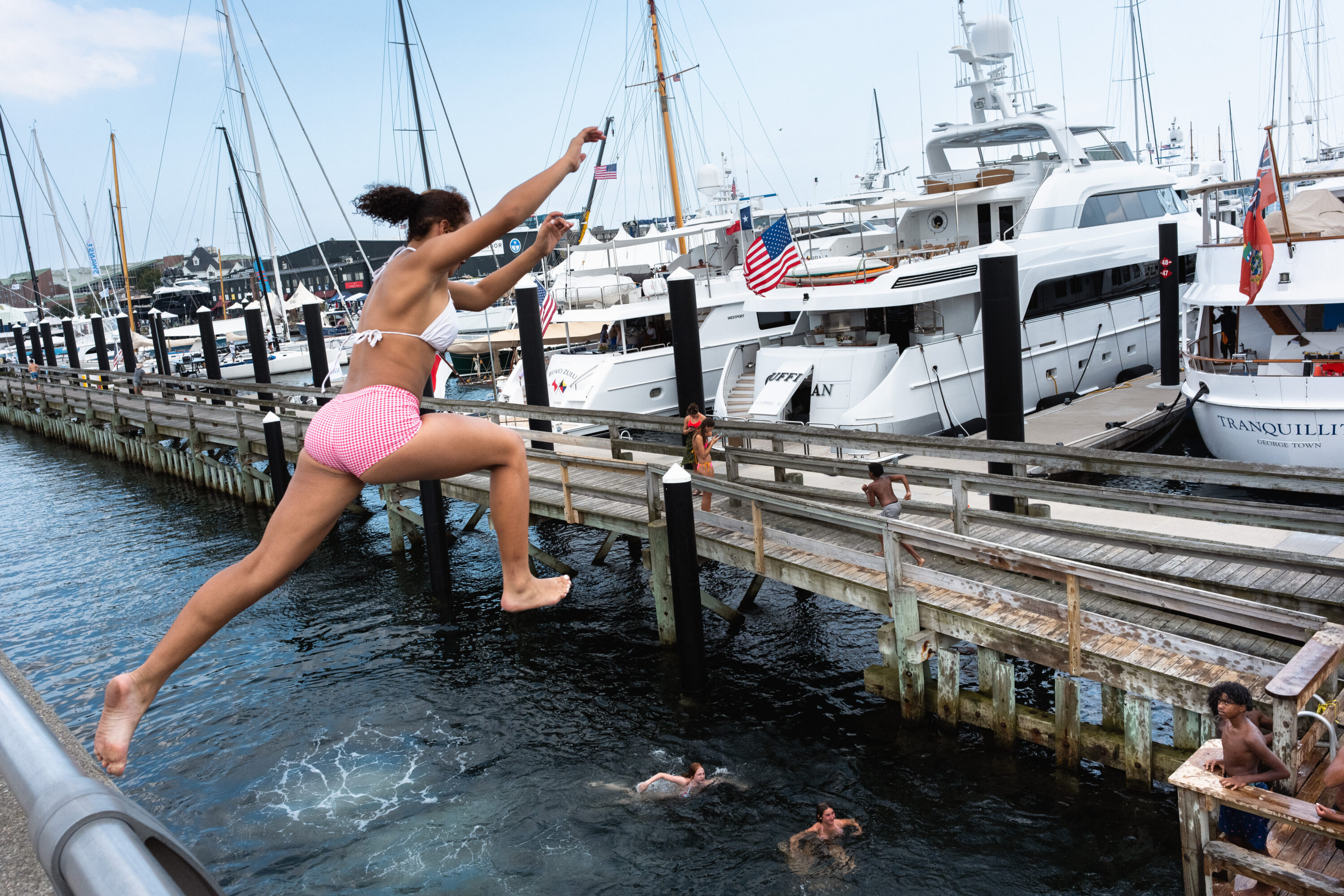 A swimmer in pink shorts leaps above the water, with other swimmers and docked yachts behind.