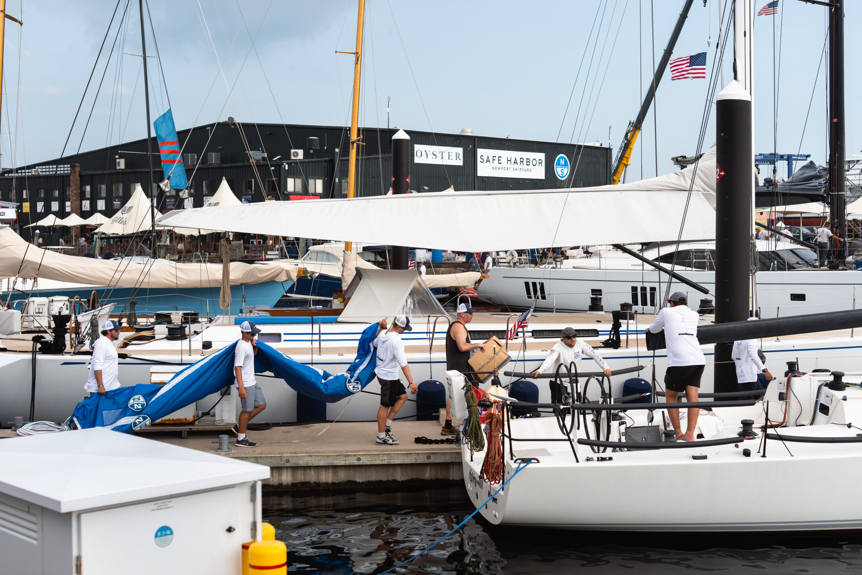 People handle a long blue sail bag on the dock beside a sailing yacht.