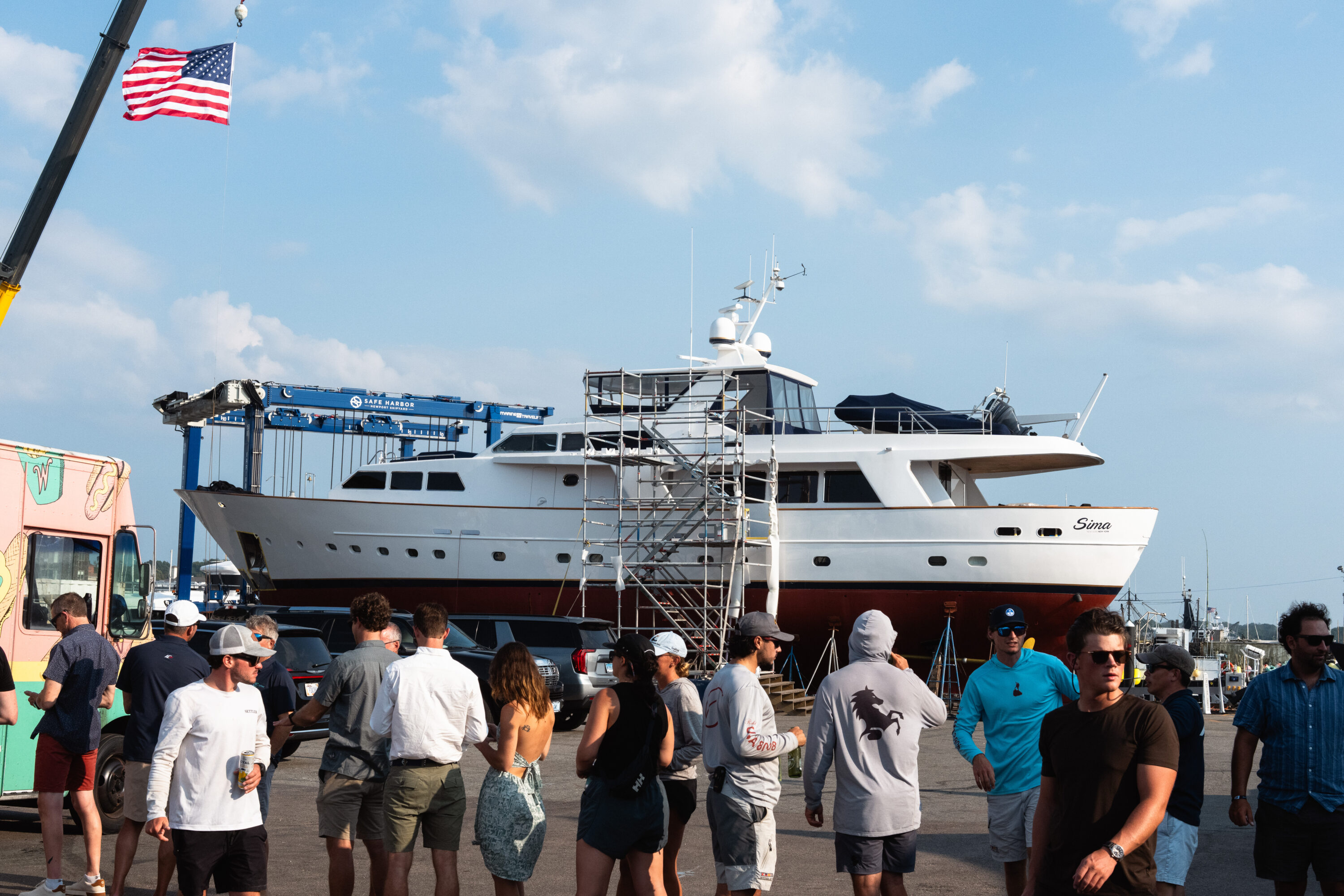A food queue crosses the foreground beneath a large yacht supported in the work yard.
