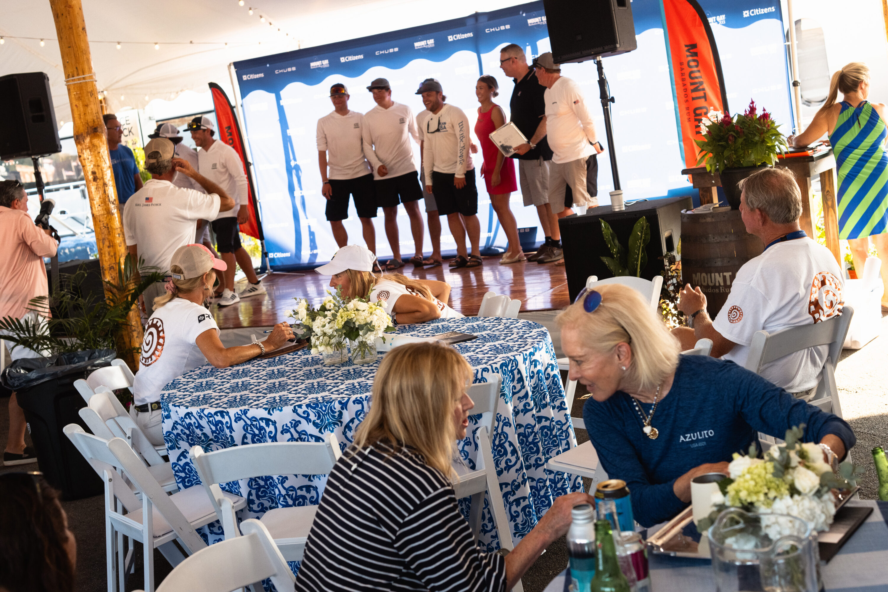 People talk at a flower-covered table while a group stands on the awards stage behind them.