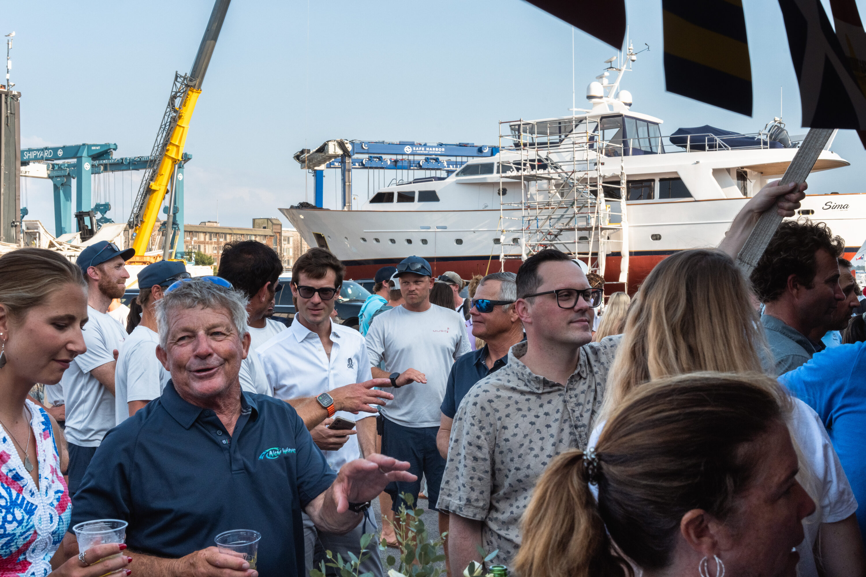 A tightly gathered crowd talks beneath a tent edge, with a yacht ashore towering behind.