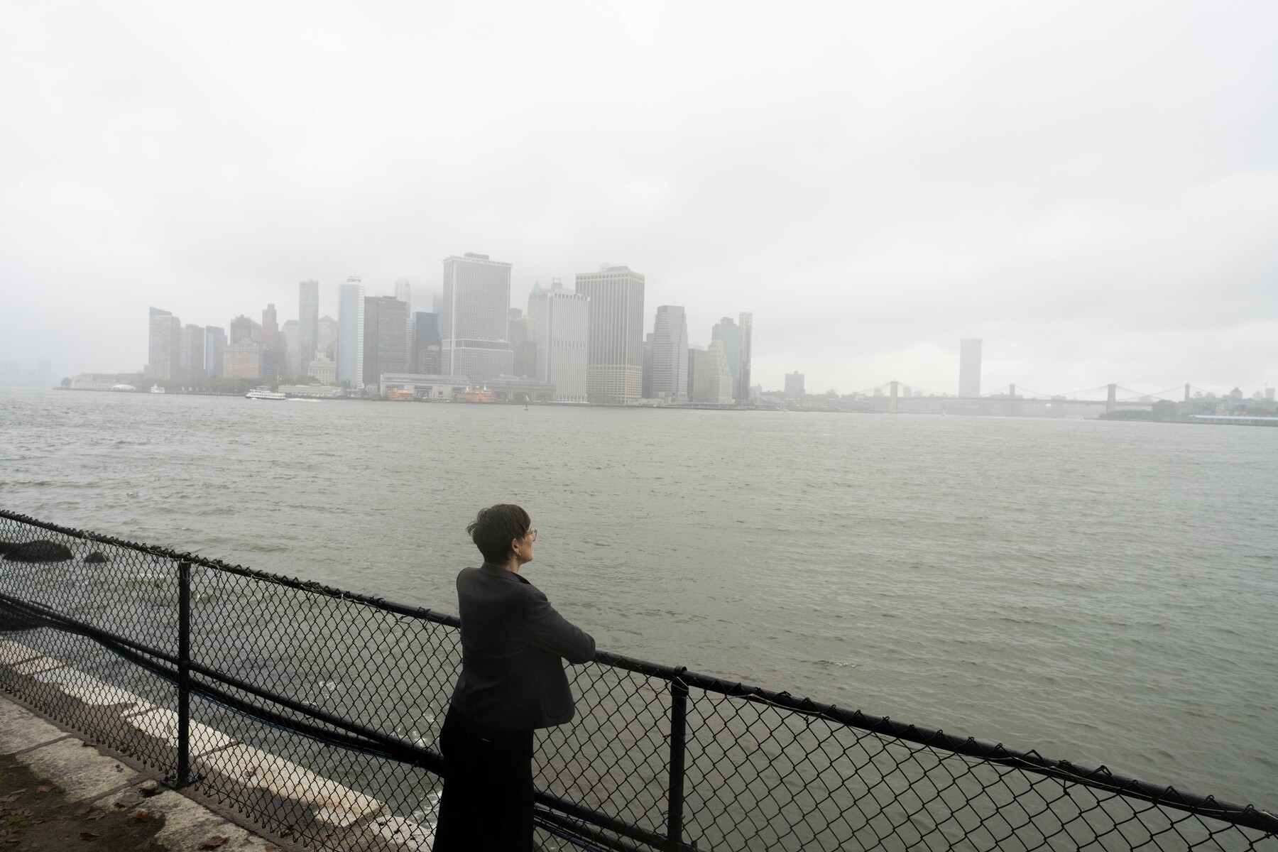A person pauses beside the Governors Island waterfront, facing a misty Lower Manhattan skyline.