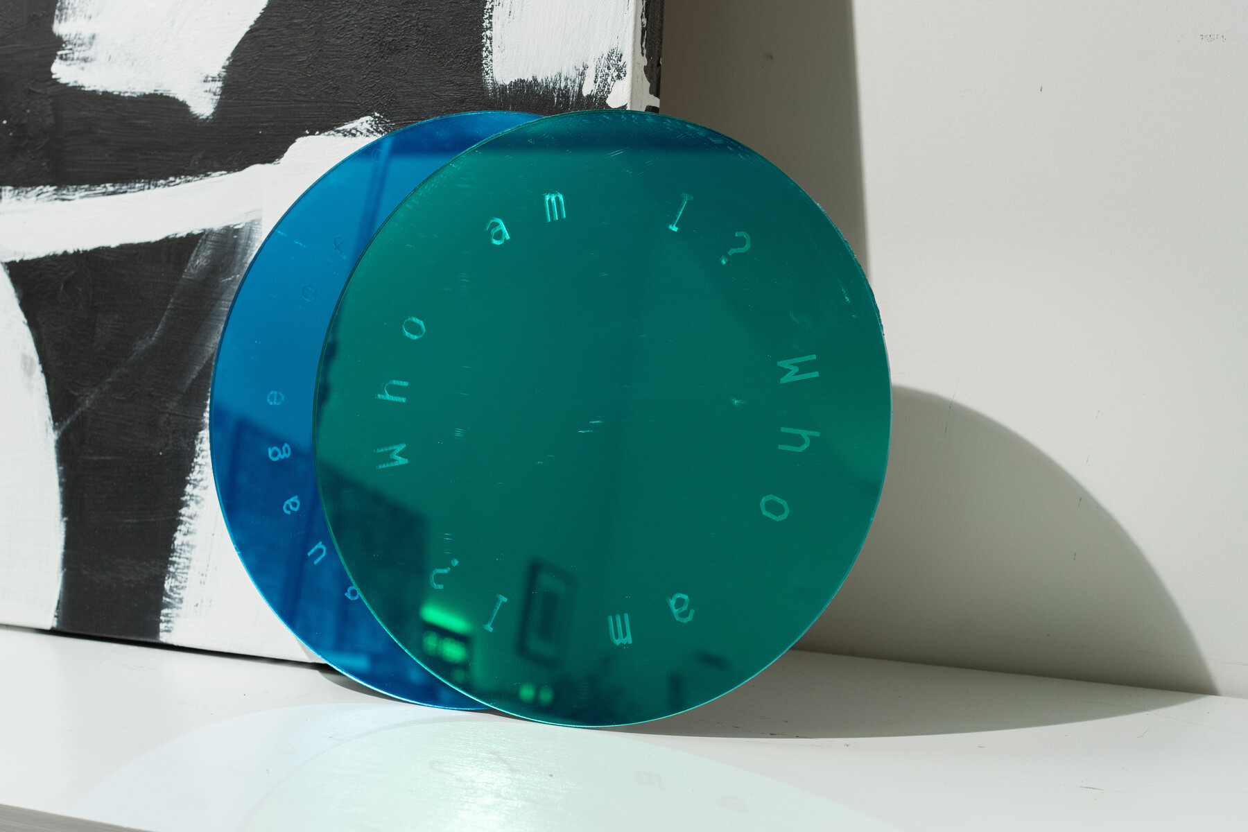 A round blue green mirror with scattered letters stands against a painted canvas.