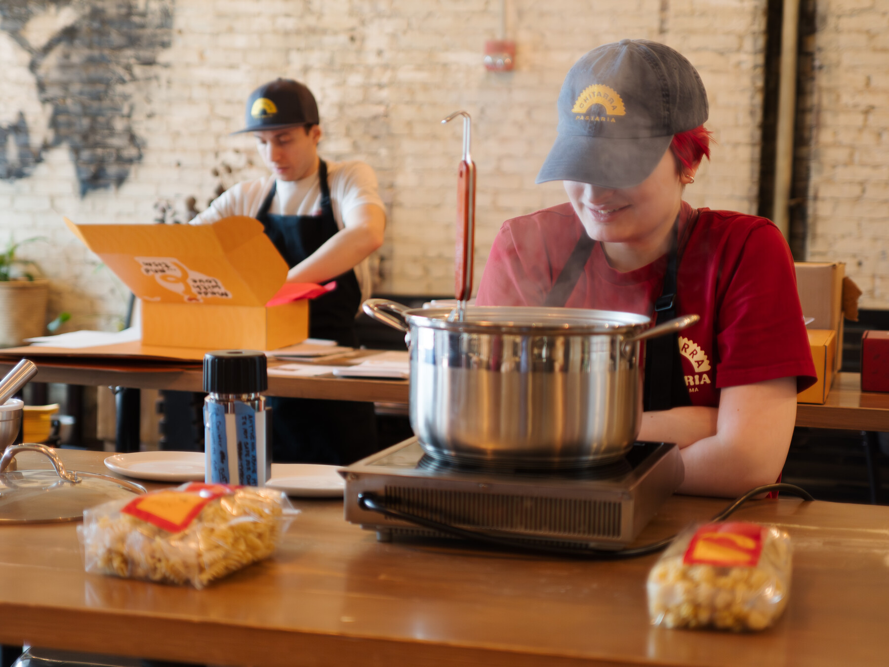 Julia Agostino tends a steaming pot while Stefano Micali packs boxes behind her.