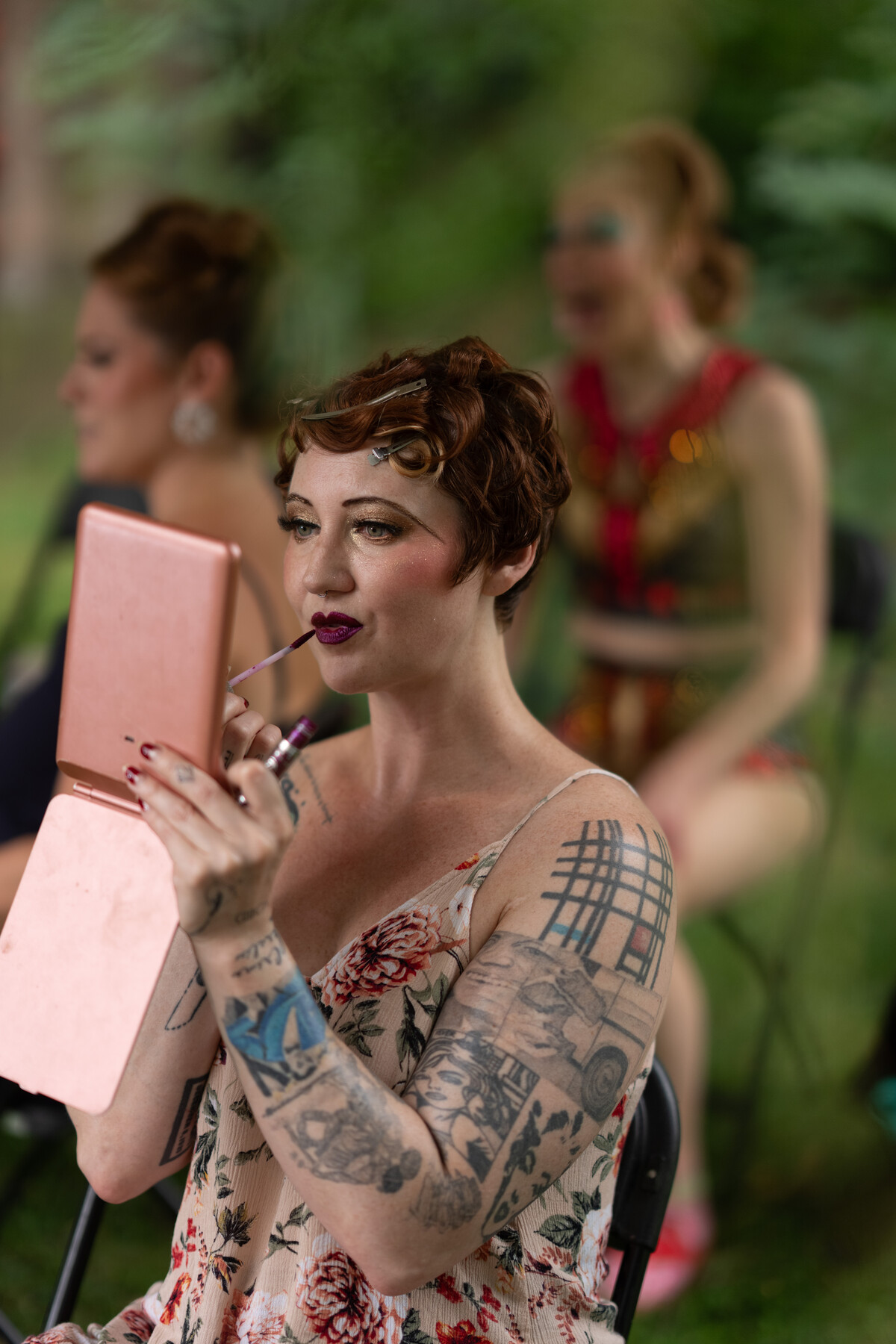 A tattooed performer applies makeup while looking into a hand mirror.
