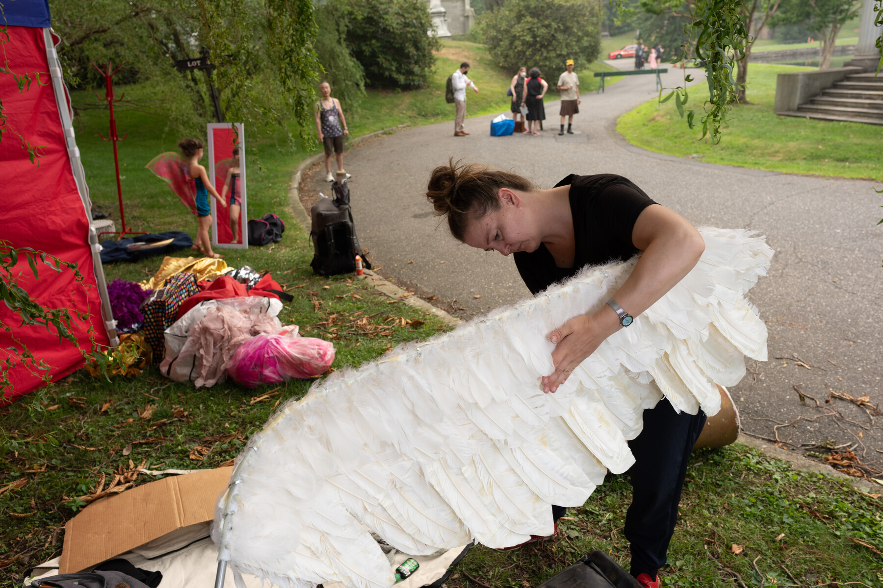 A performer prepares a large white feathered wing beside a cemetery path.