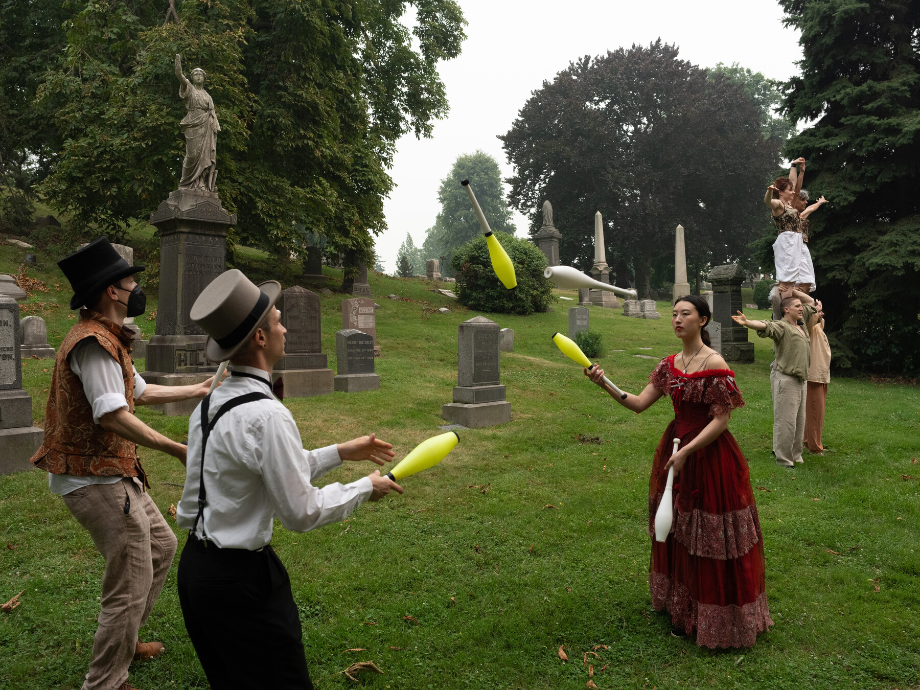 Costumed performers juggle clubs in a grassy cemetery clearing.