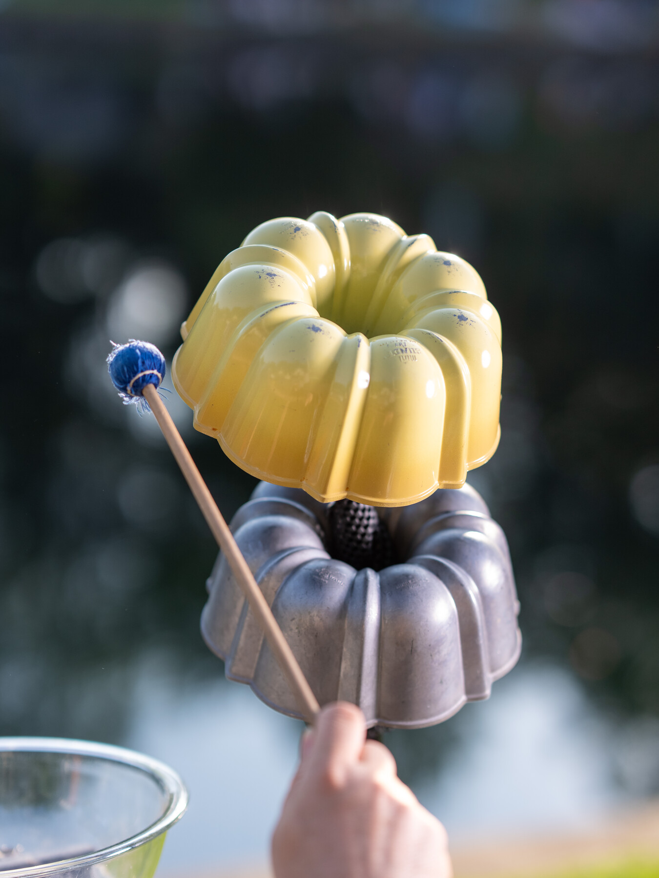 A hand holds a mallet beside a bright yellow metal mold used as percussion.