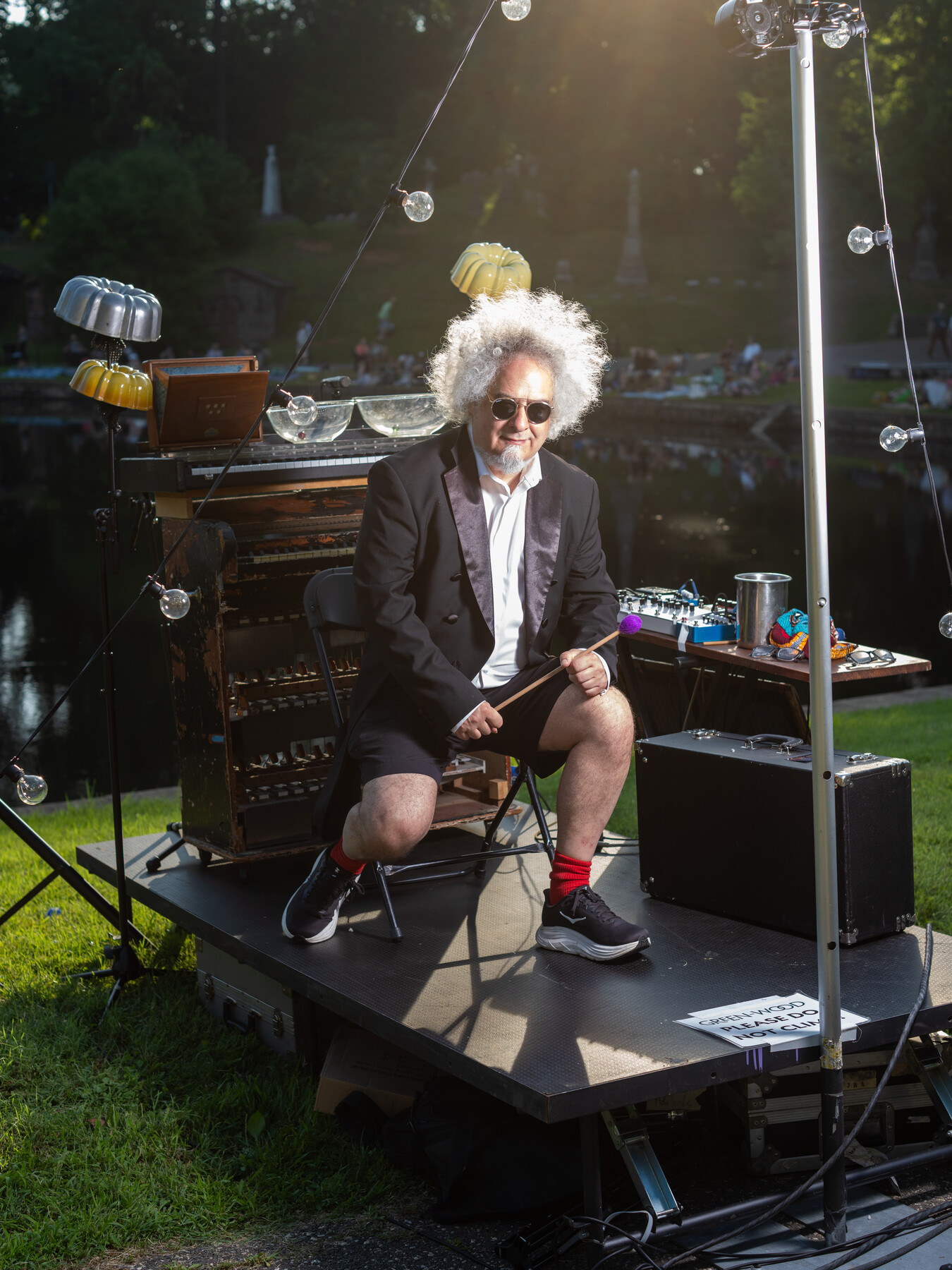A gray-haired musician sits on a small outdoor stage with instruments behind him.
