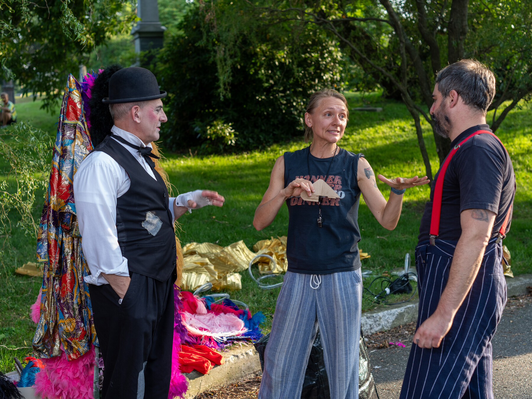 Three performers talk beside colorful costumes spread beneath trees.