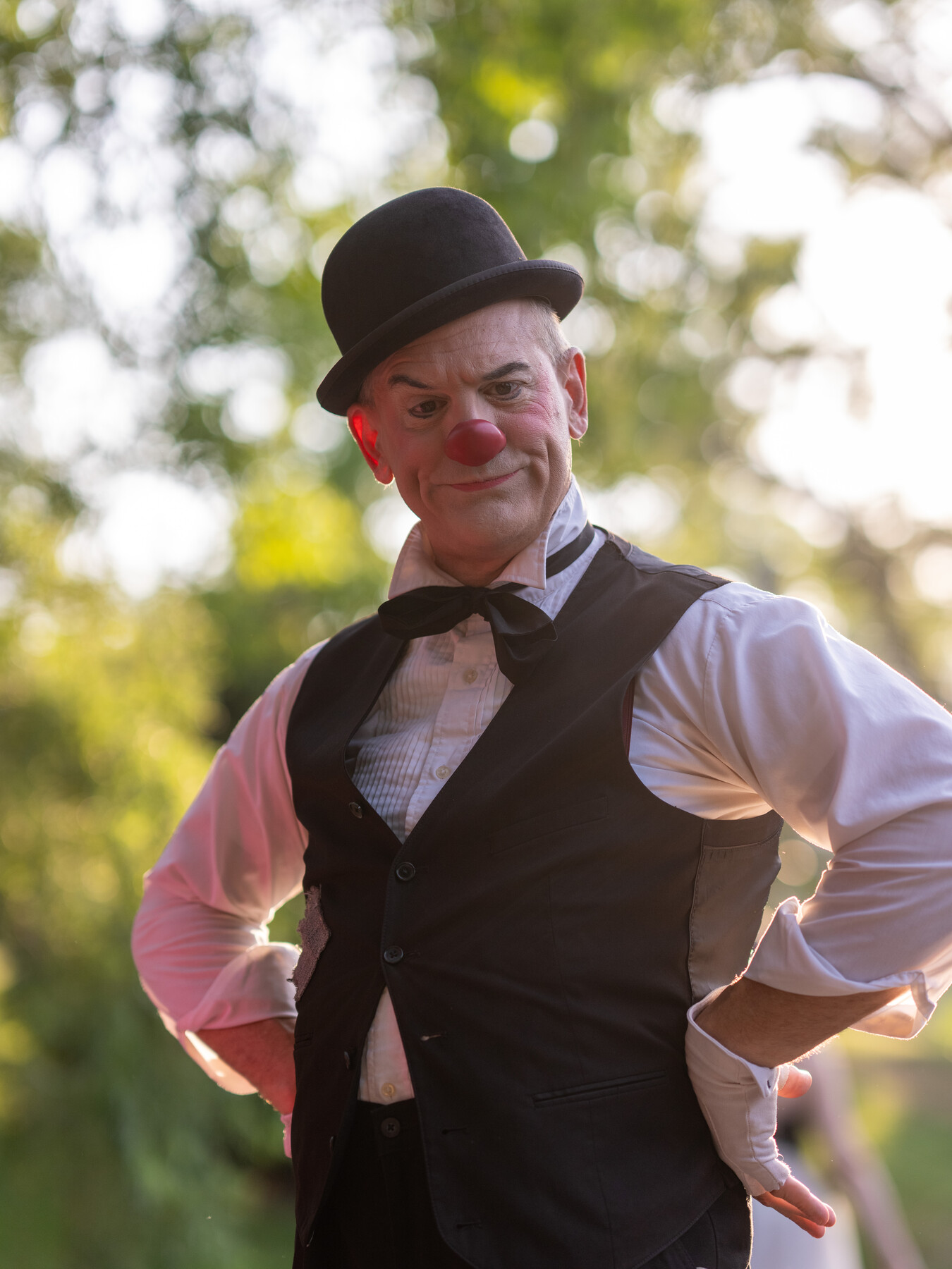 A clown in a bowler hat and red nose smiles outdoors.