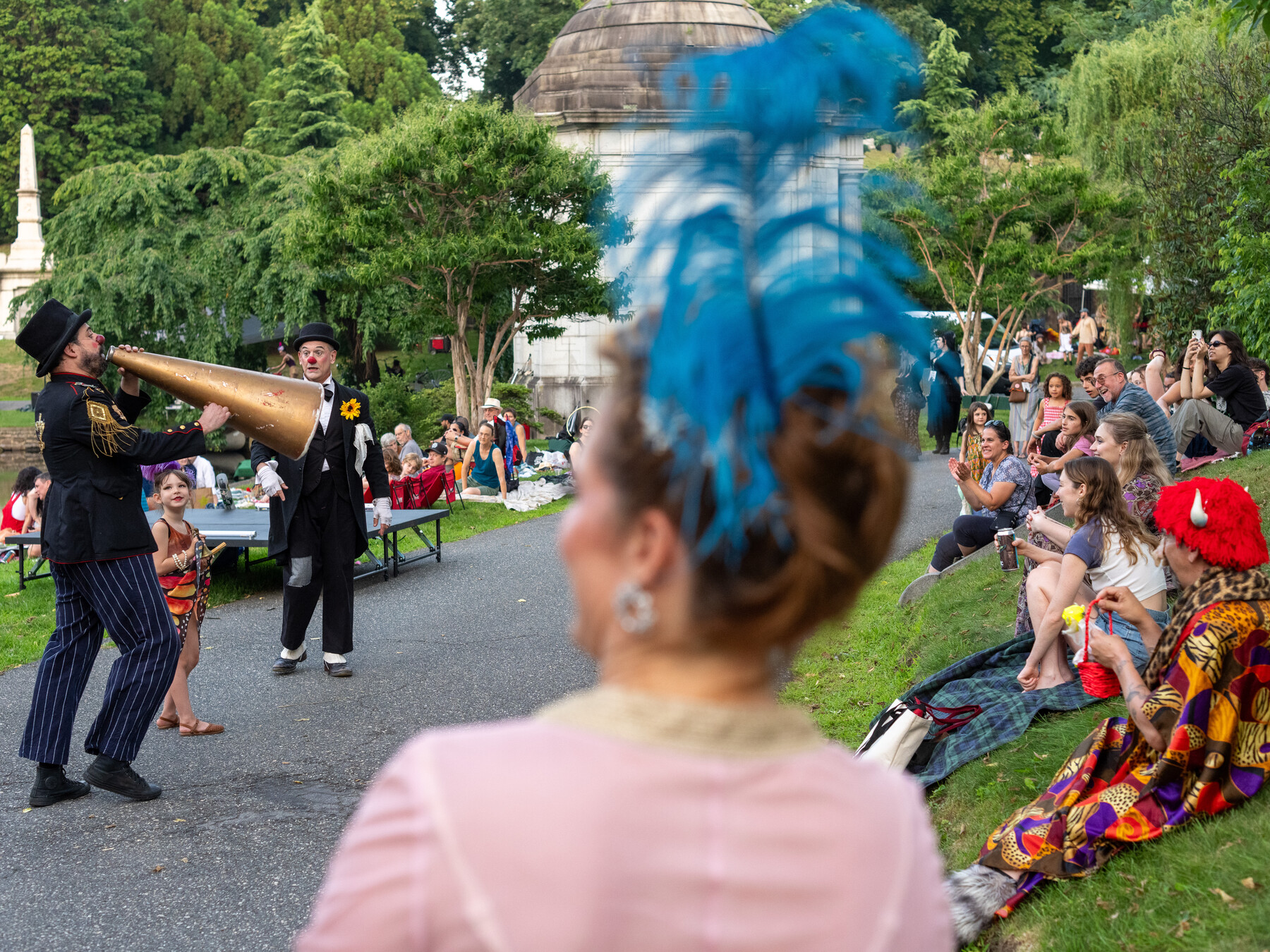 Performers use long horns among spectators gathered on a cemetery path.
