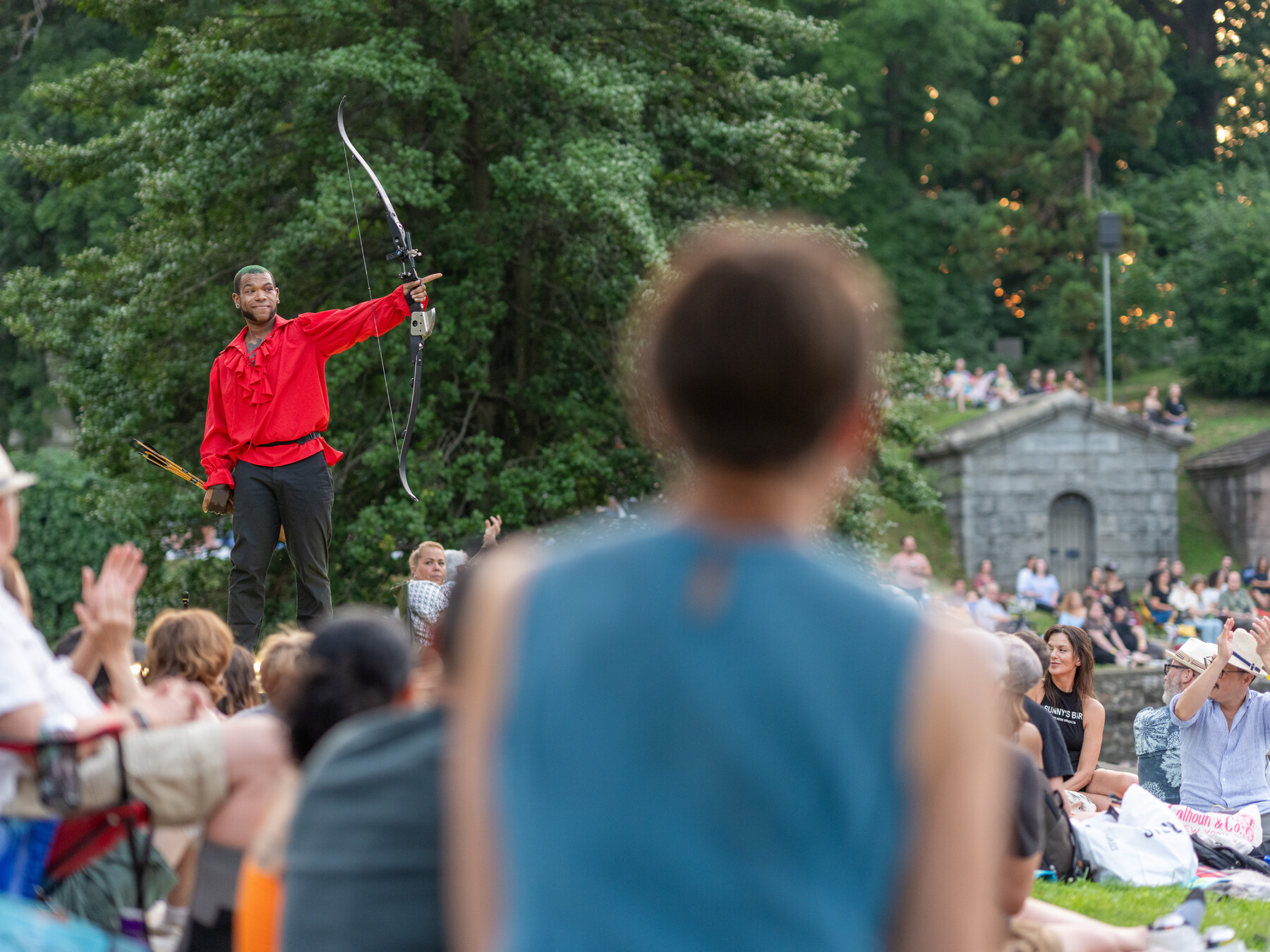 A performer in a red shirt raises a hoop while the crowd watches near the lake.