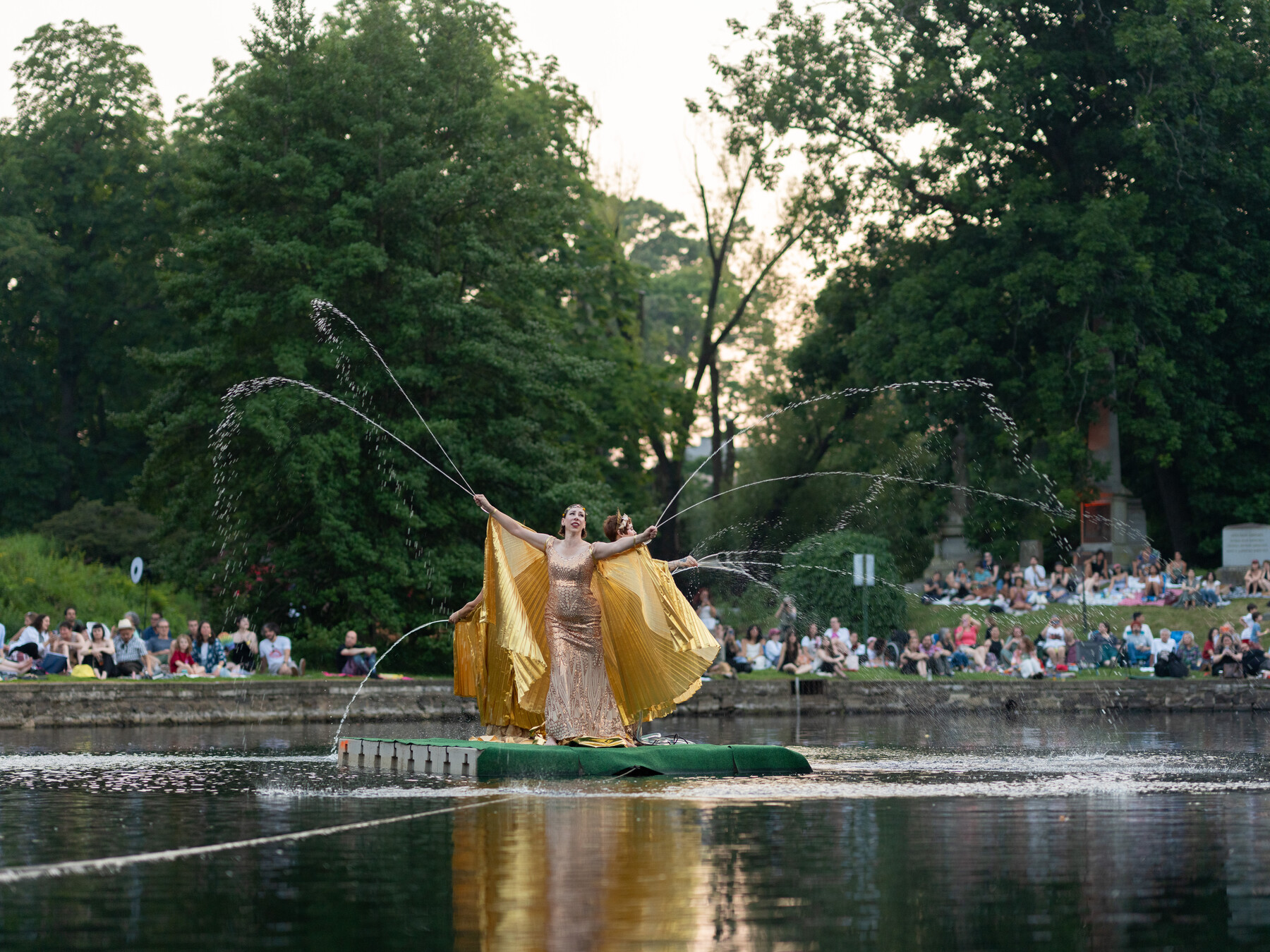 A gold-costumed performer dances amid water jets on a floating platform.