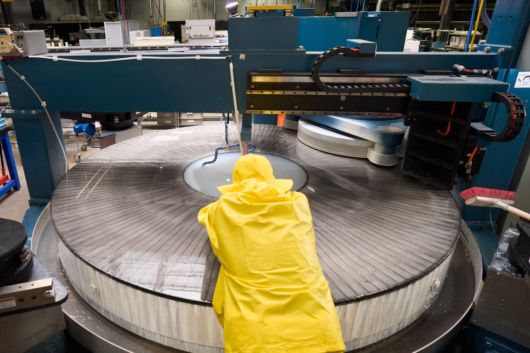 A person in a bright yellow hooded coat faces a very large circular machine surface at Optimax in Ontario, New York.