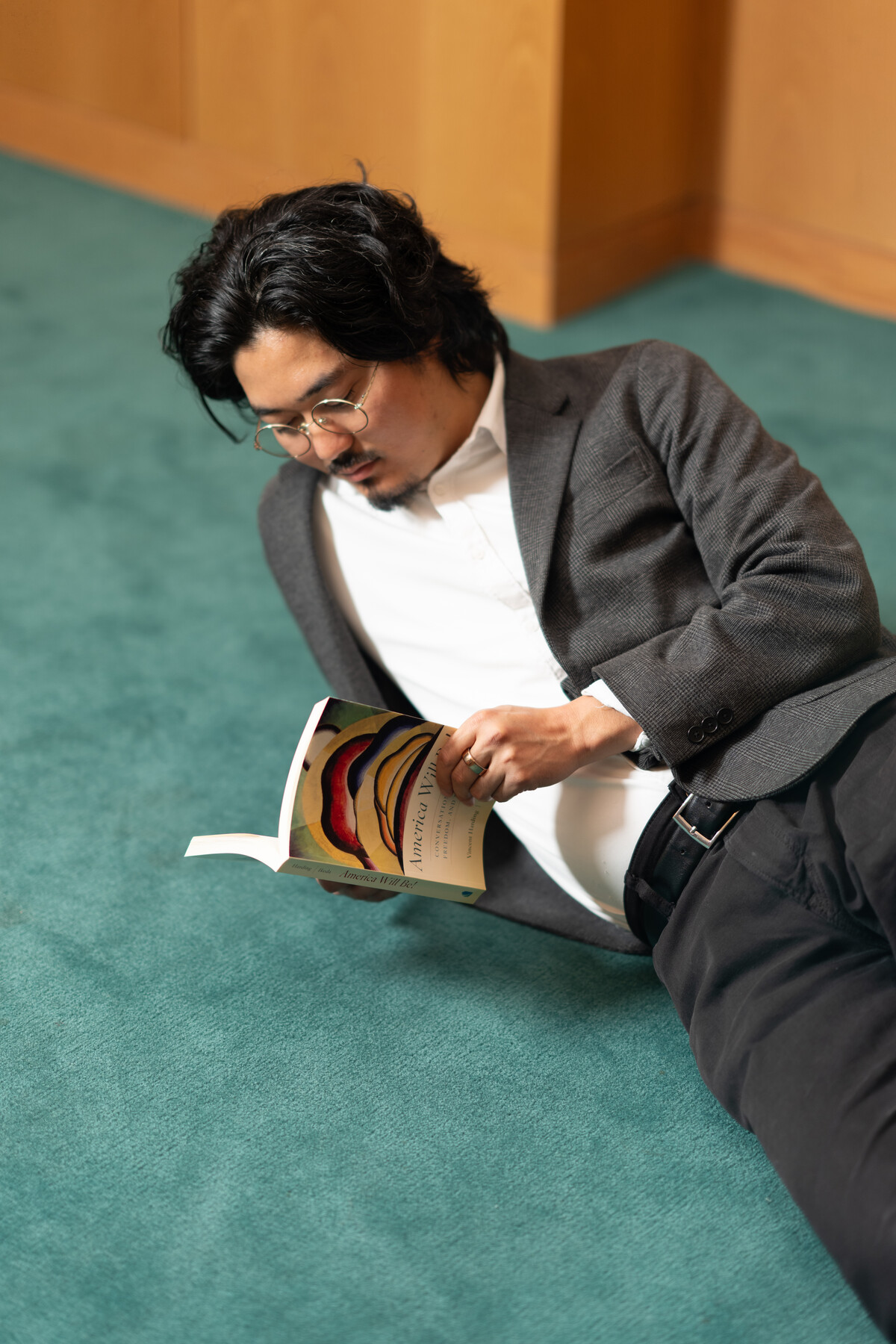 Staff member reclining on teal carpet and reading America Will Be! beside wooden cabinetry.