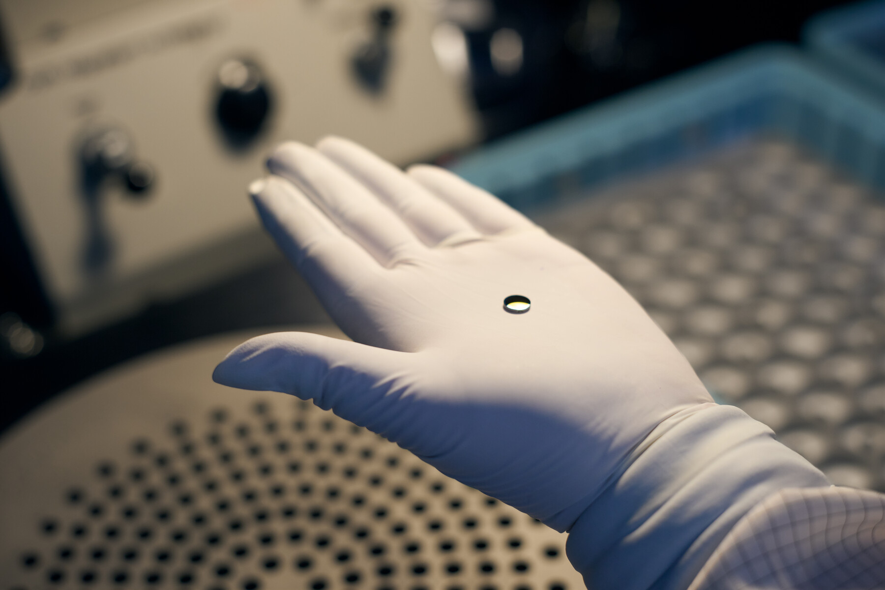 A very small round reflective object rests alone on an open white-gloved palm at Optimax in Ontario, New York.