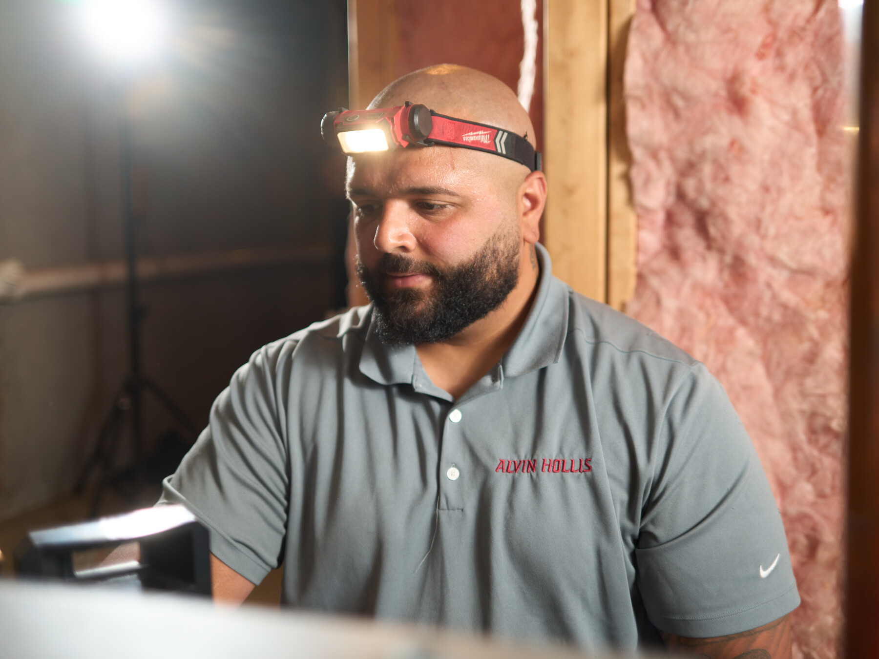 A bearded technician in an Alvin Hollis polo wears a lit headlamp in a basement.