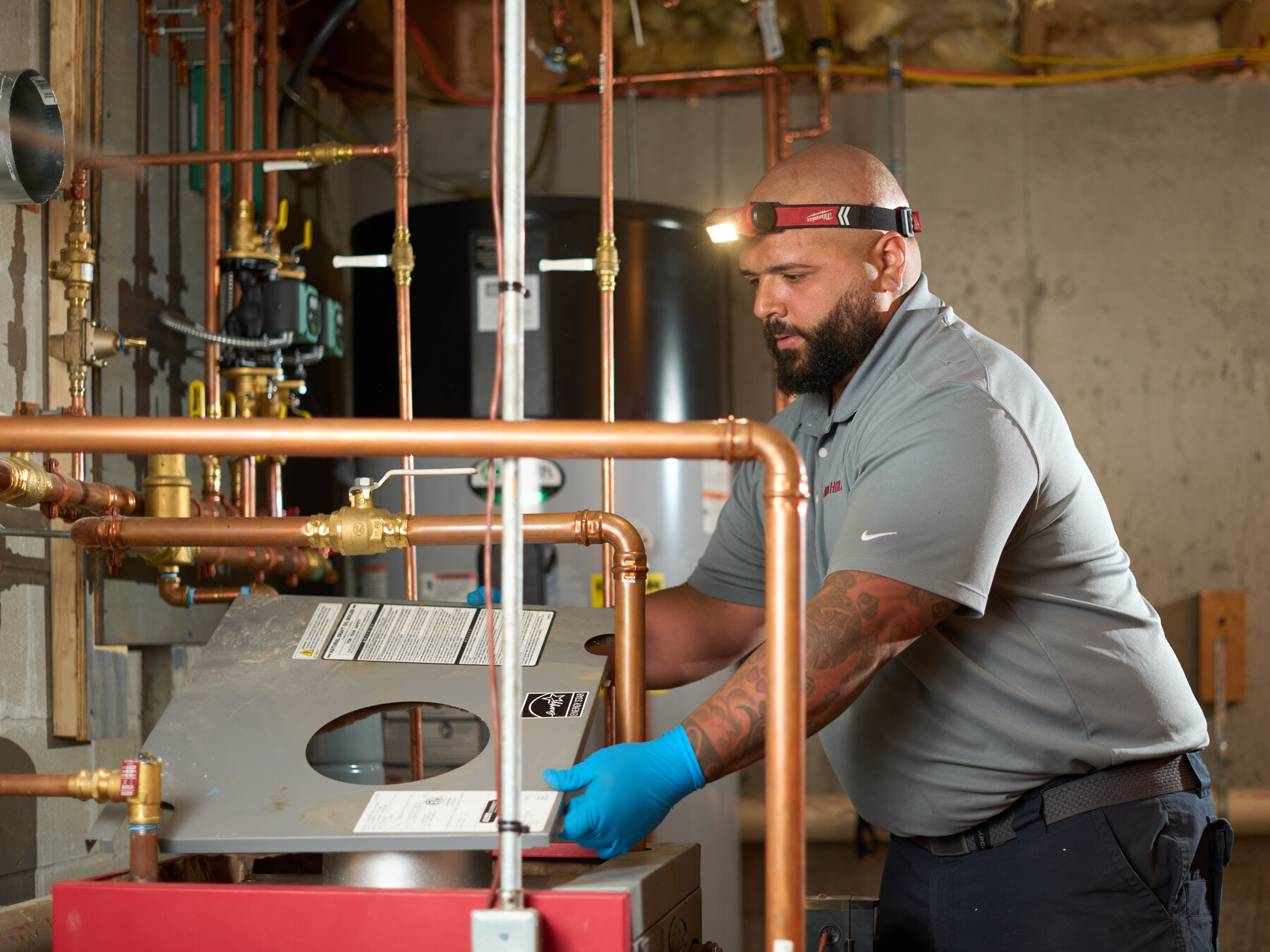 An Alvin Hollis technician wearing a headlamp lifts a heating-unit cover among copper pipes.