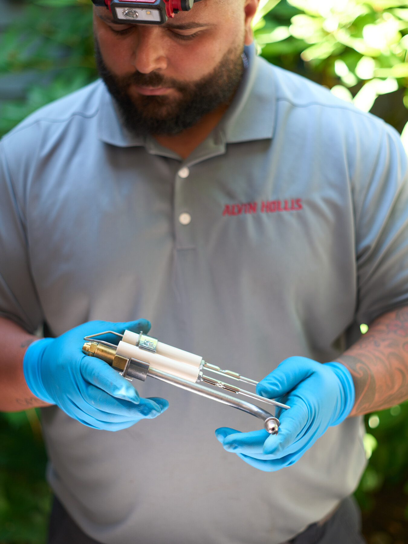 A technician holds a narrow metal component between blue-gloved hands outdoors.