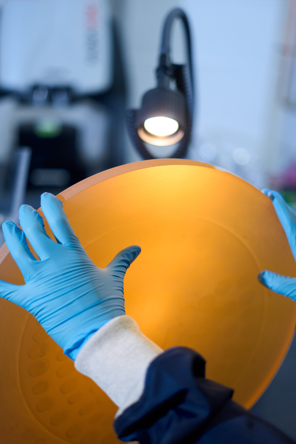 Blue-gloved hands hold a large amber-colored circular object beneath a lamp at Optimax in Ontario, New York.