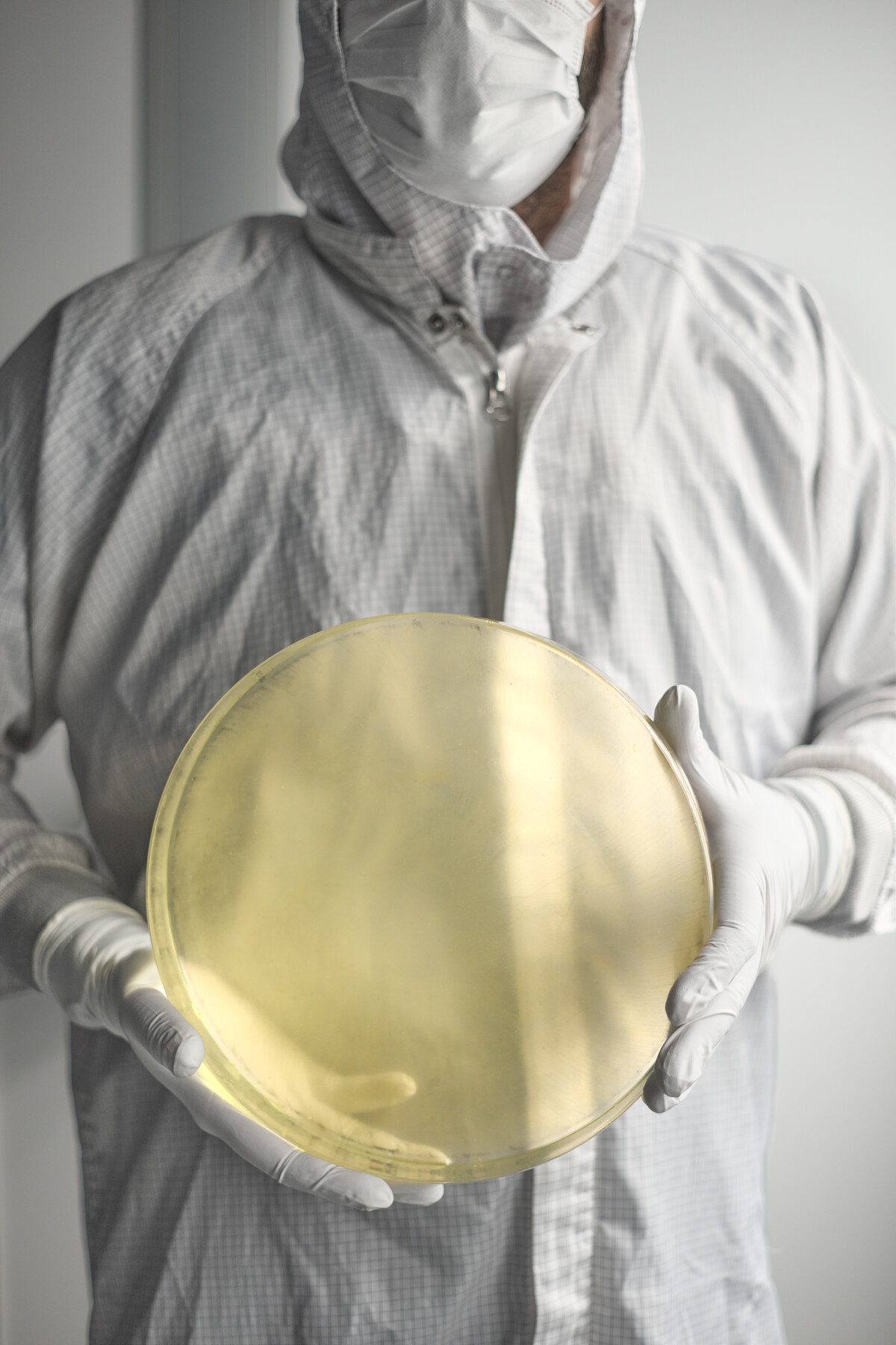 A person in a mask and protective clothing holds a large gold-colored circular object with white-gloved hands at Optimax in Ontario, New York.