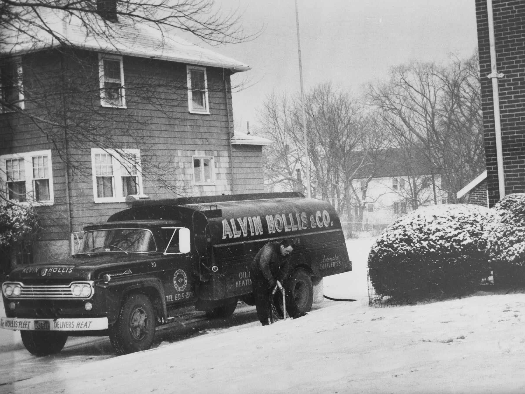 An undated black-and-white photograph of an Alvin Hollis fuel truck and a worker beside a snow-covered house.
