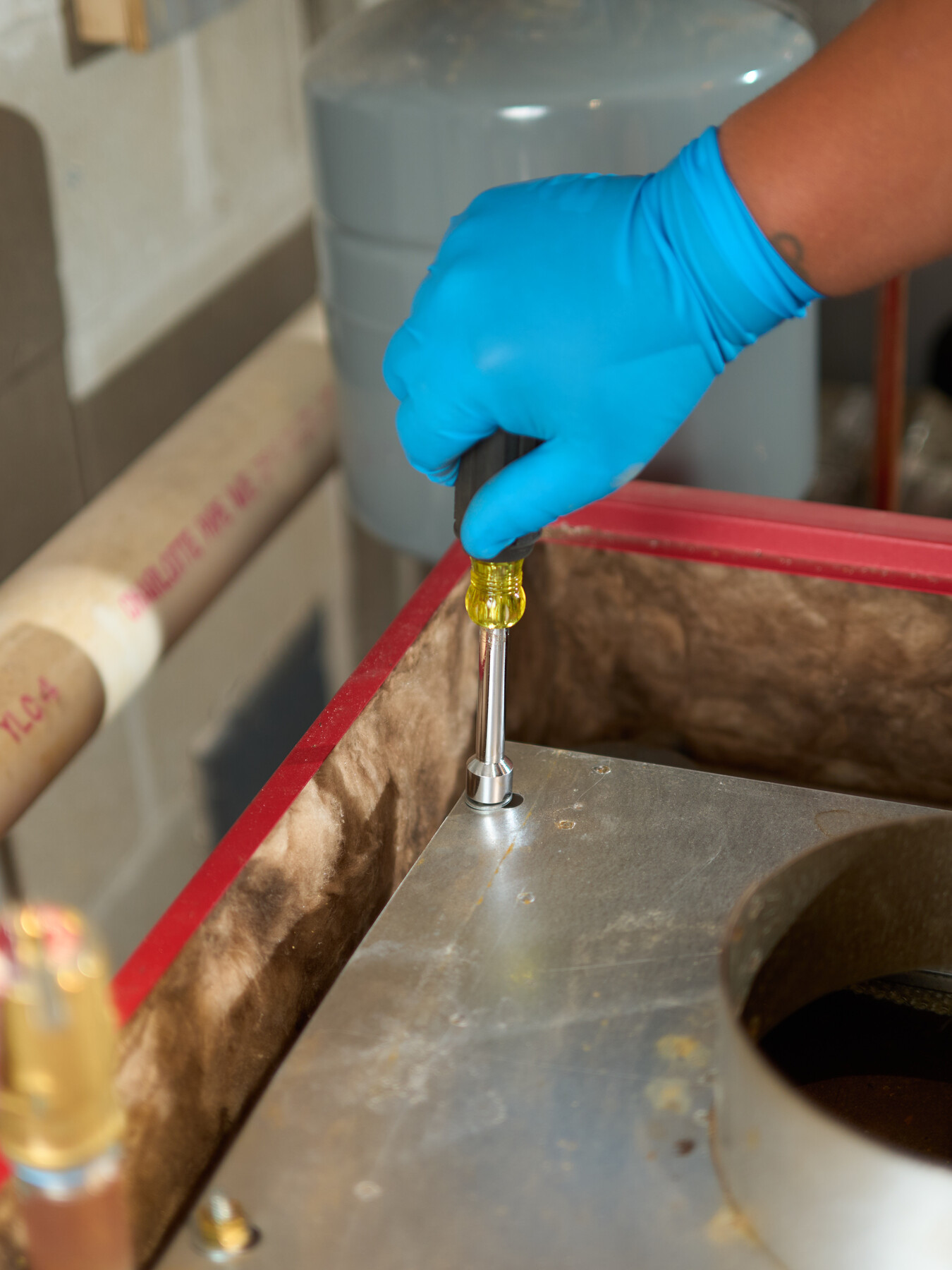 A blue-gloved hand turns a nut driver on the metal cover of a red heating unit.