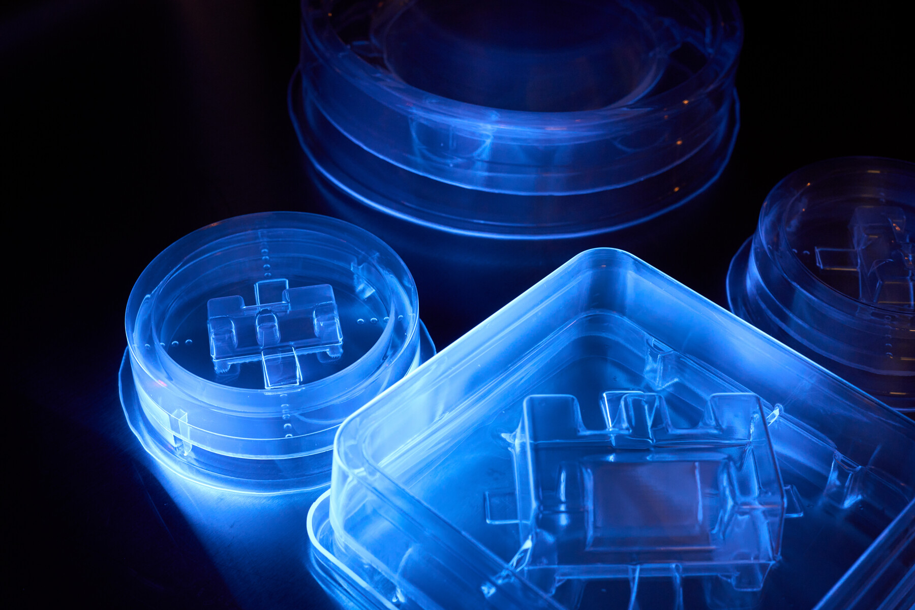 Transparent containers and small clear pieces glow blue against a dark background at Optimax in Ontario, New York.
