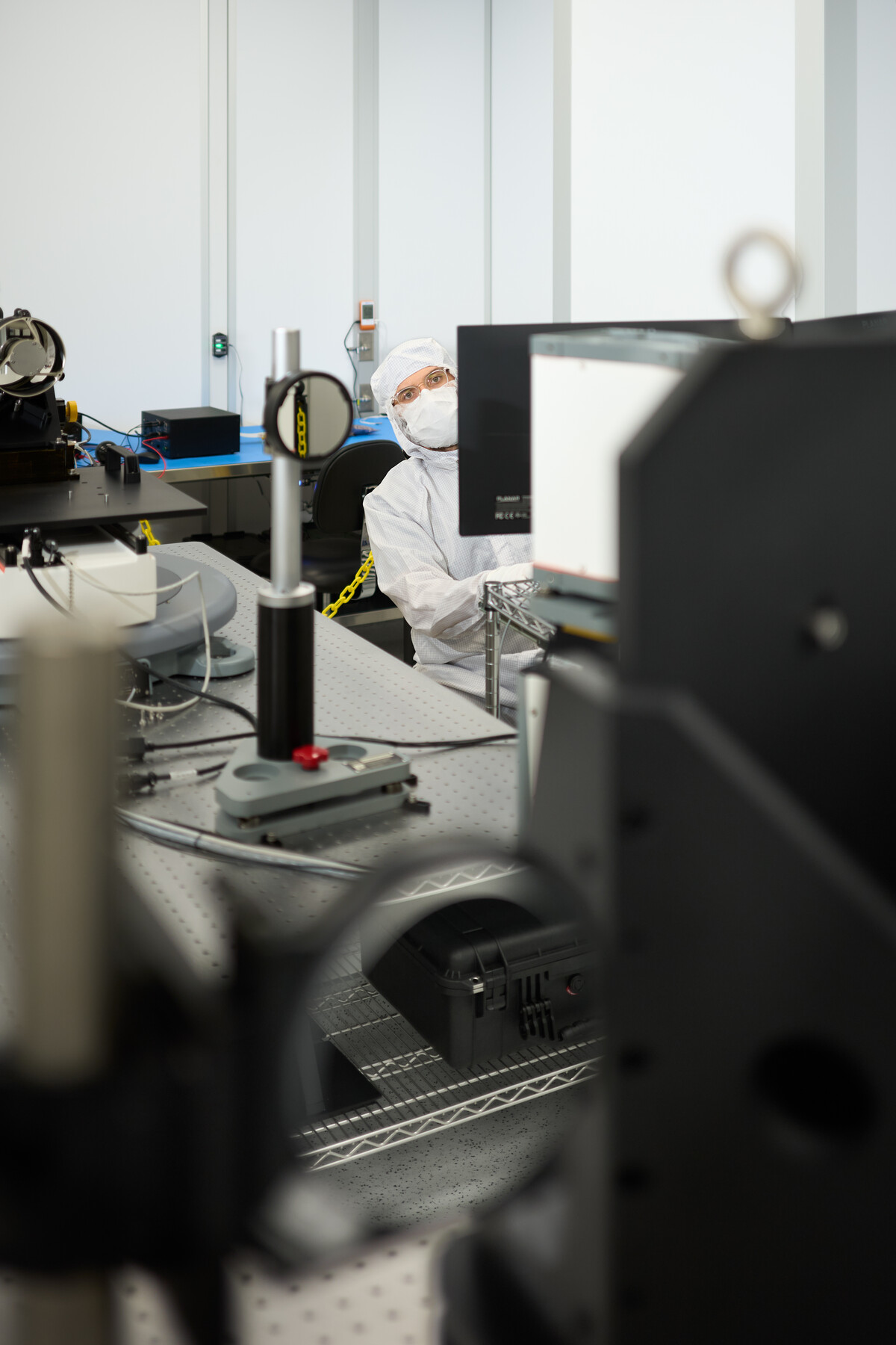 A person wearing glasses, a mask and protective clothing peeks around a computer monitor amid optical equipment at Optimax in Ontario, New York.