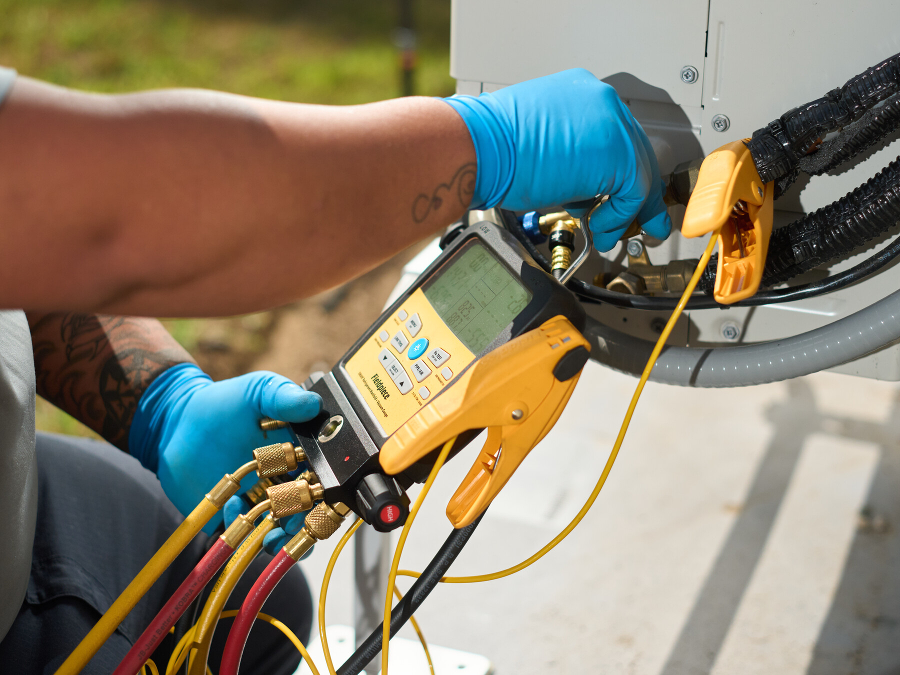 Blue-gloved hands handle a yellow diagnostic manifold and hoses connected to an outdoor unit.