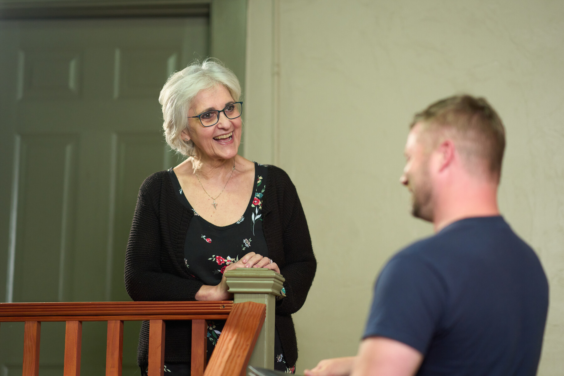 A woman smiles toward a man in a navy shirt beside an indoor wooden railing.