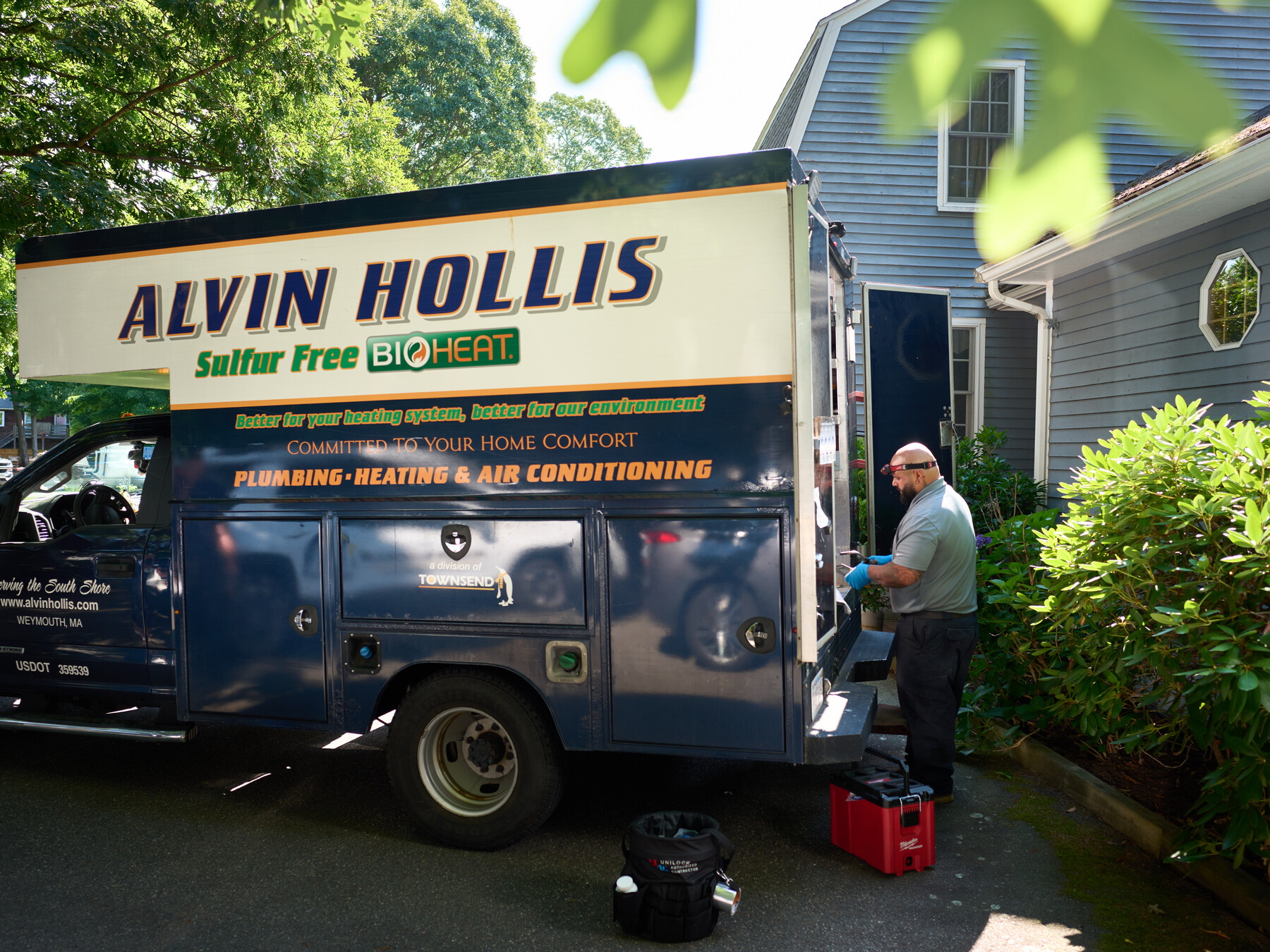 An Alvin Hollis service truck stands beside a blue house as a technician works at its open rear.