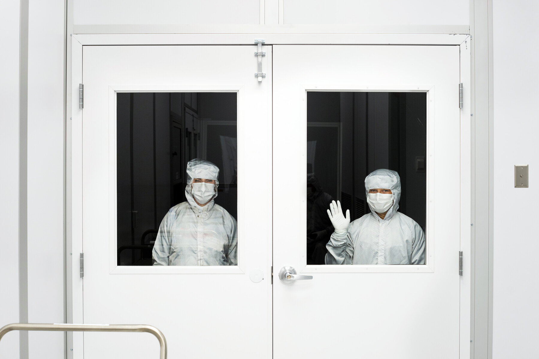 Two people in protective clothing look through separate door windows; one raises a gloved hand at Optimax in Ontario, New York.