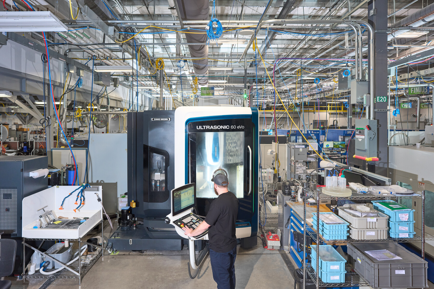 A person operates a machine in a wide factory interior filled with pipes, cables and trays at Optimax in Ontario, New York.