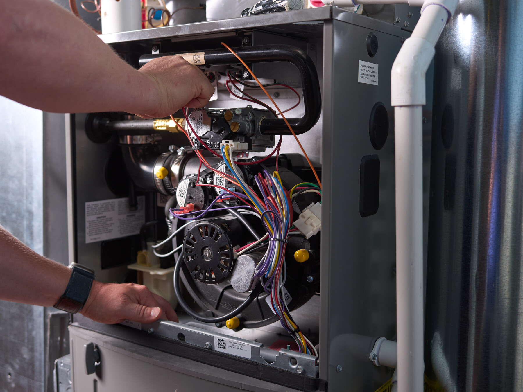 Hands reach into the open casing of a heating unit among wiring, pipes and mechanical parts.