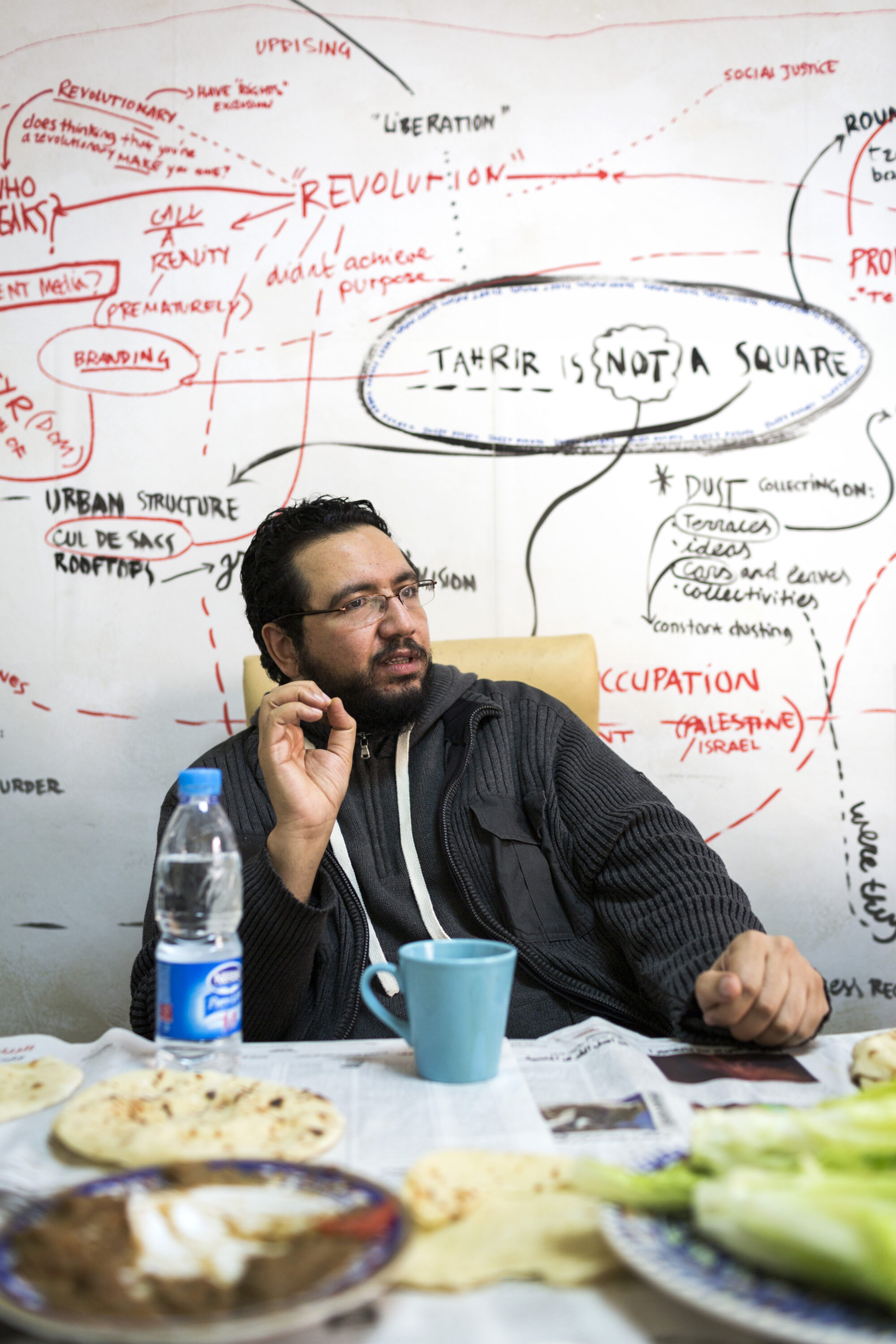 Belal Fadl gestures at a table with bread, vegetables and a turquoise cup in Mada Masr’s newsroom.