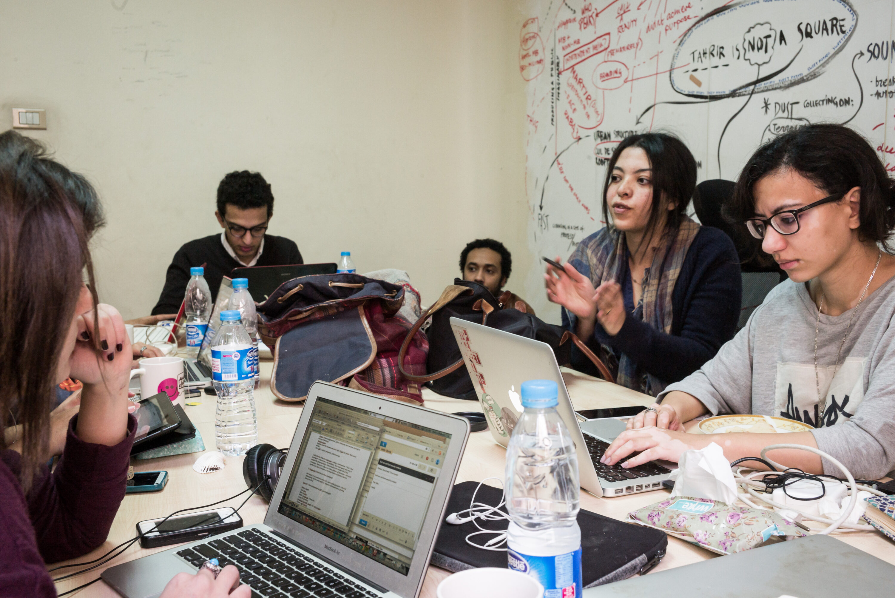 Mada Masr journalists gather around a table of laptops beneath a handwritten wall diagram.