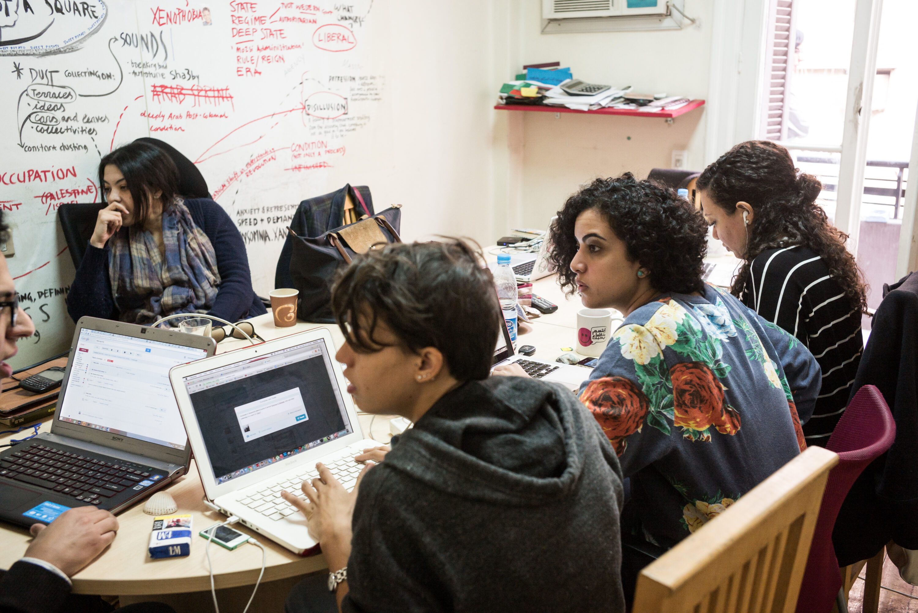 Journalists work and talk around a crowded table of laptops in Mada Masr’s Cairo newsroom.