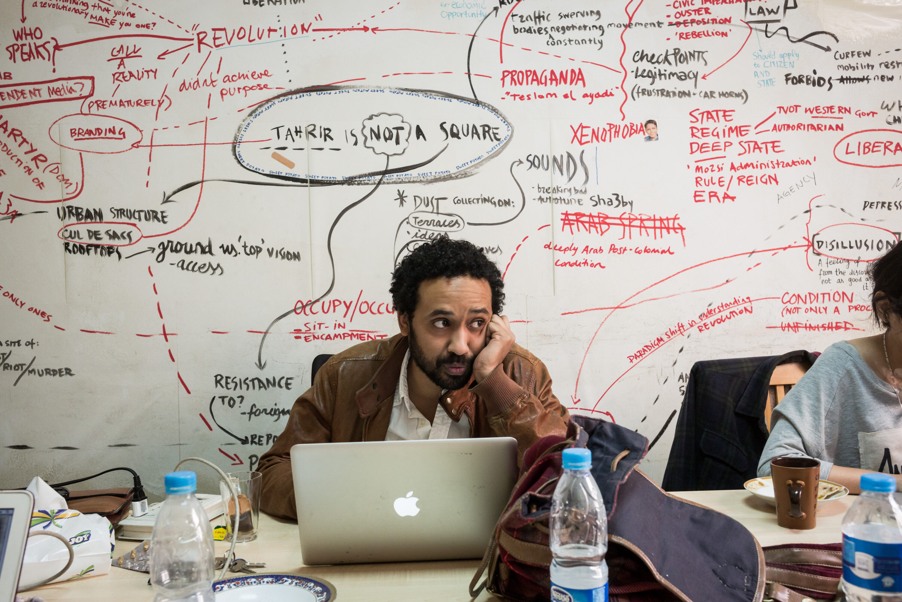 A Mada Masr editor sits behind a laptop with his cheek resting on one hand, beneath a handwritten wall diagram.