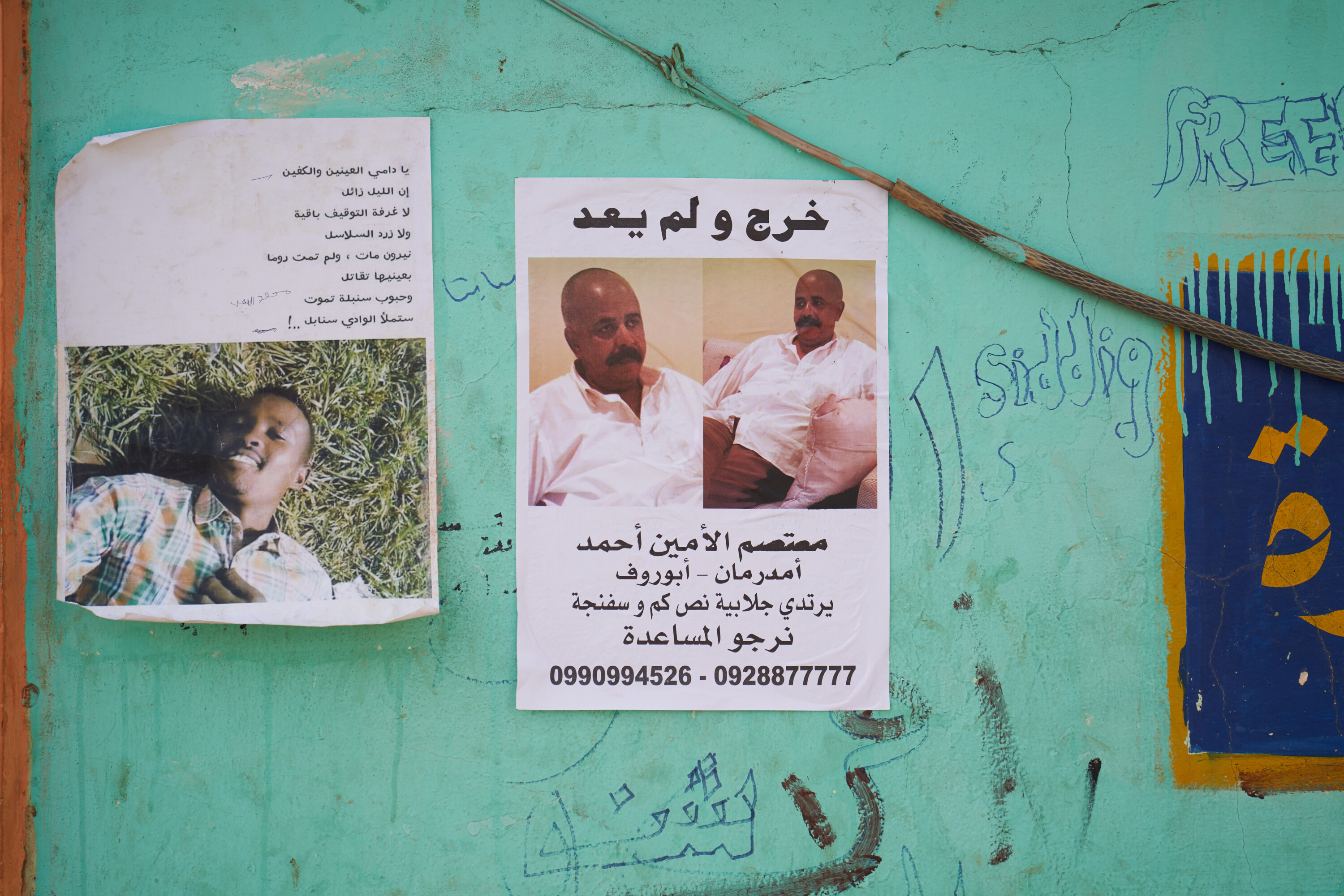 Two posters with portraits and Arabic text hang on a turquoise wall covered in handwritten protest slogans.