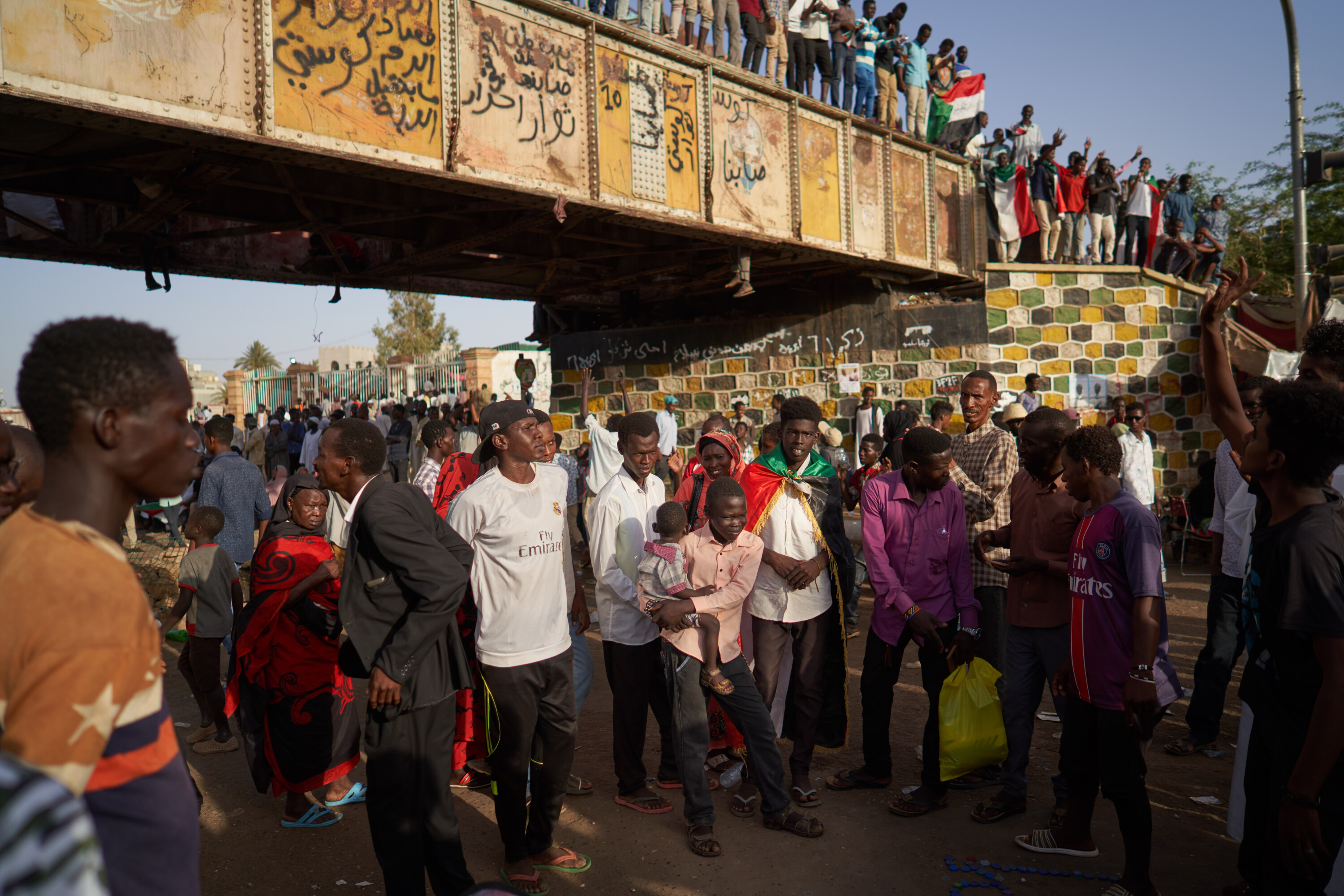Protesters gather beneath a graffiti-covered railway bridge while others stand above them holding Sudanese flags.