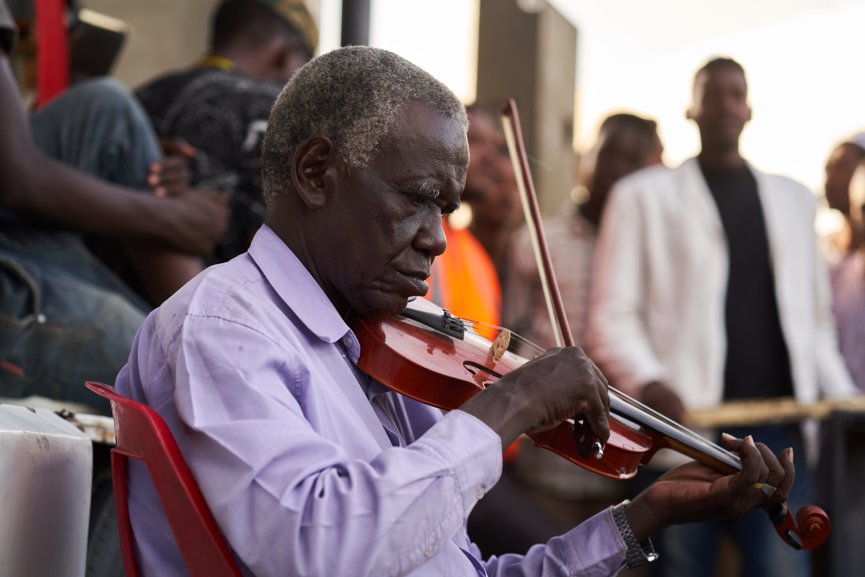 A seated man in a pale purple shirt plays a violin, with people gathered behind him at the Khartoum sit-in.