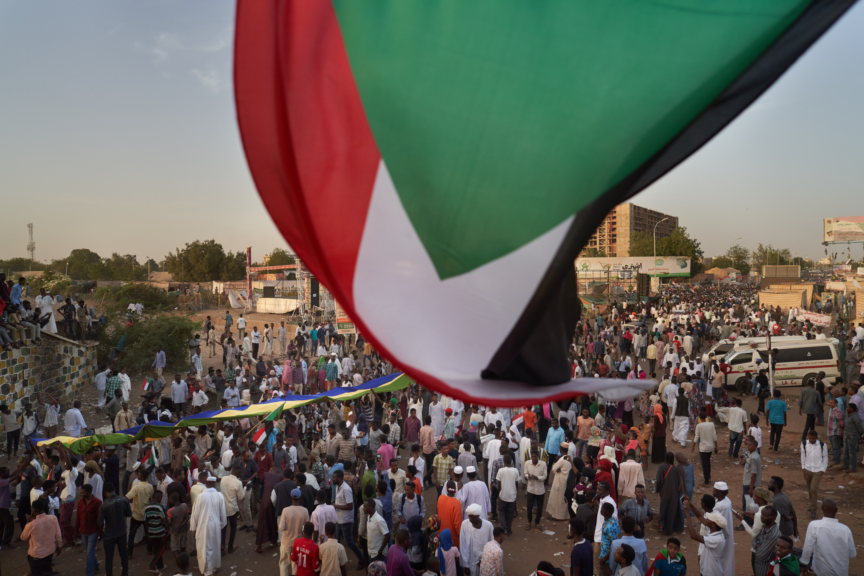 A large Sudanese flag sweeps across the foreground above a crowd walking among tents and banners at the sit-in.